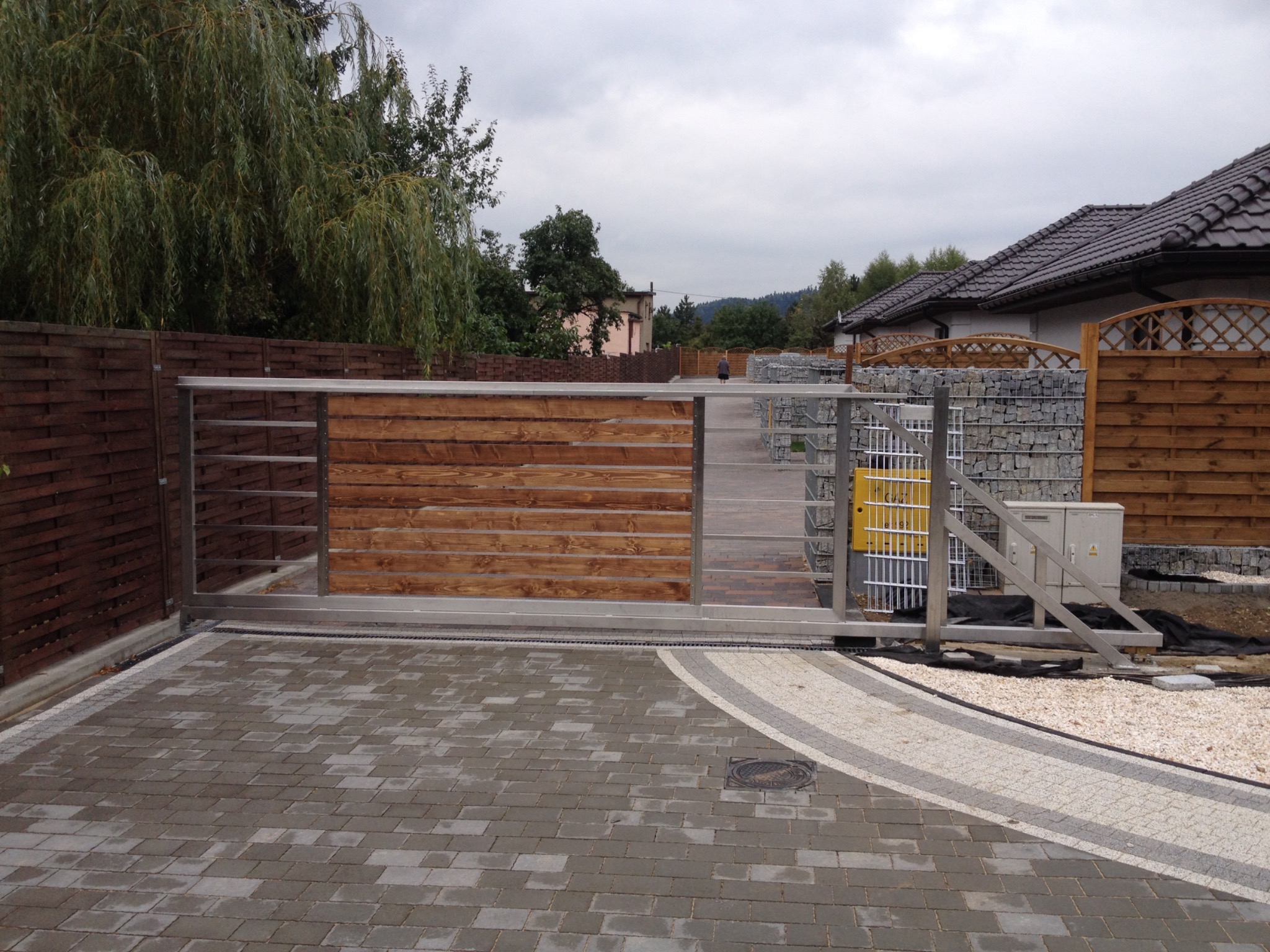 Suwana brama z drewnianymi panelami w stalowej ramie, widok z zewnątrz na posesję wyłożoną kostką brukową, z elementami ogrodzenia z gabionów w tle.