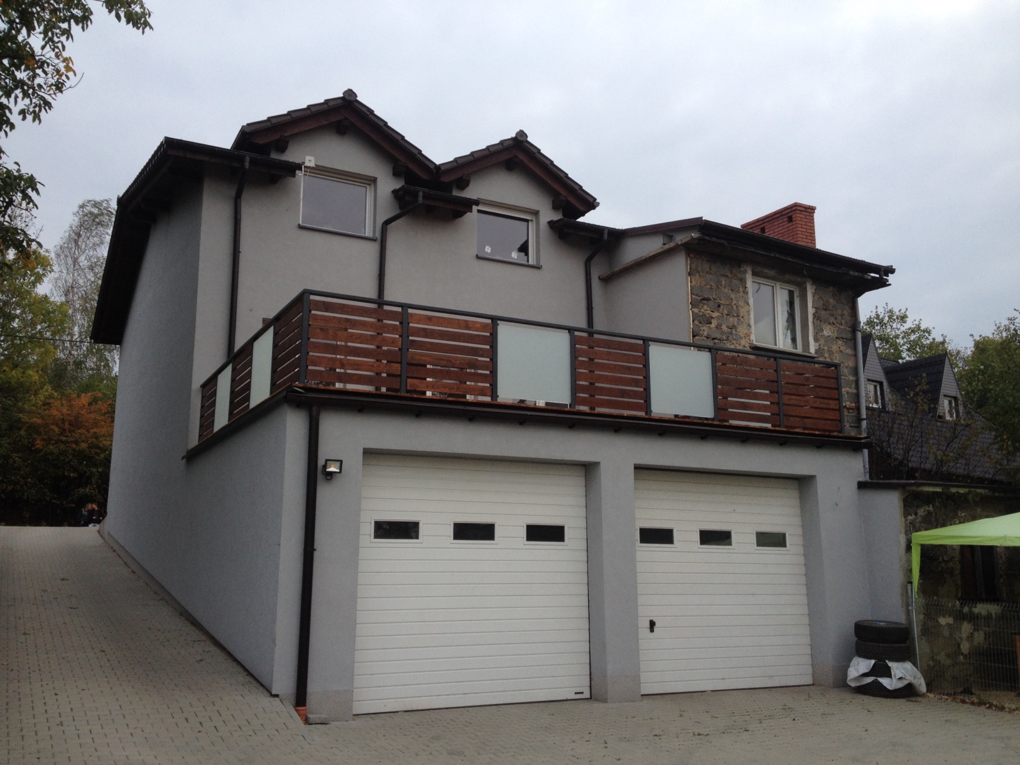 Szary dom z drewniano-szklaną balustradą balkonu nad dwoma białymi bramami garażowymi, widok z dołu na pochylony podjazd z kostki.
