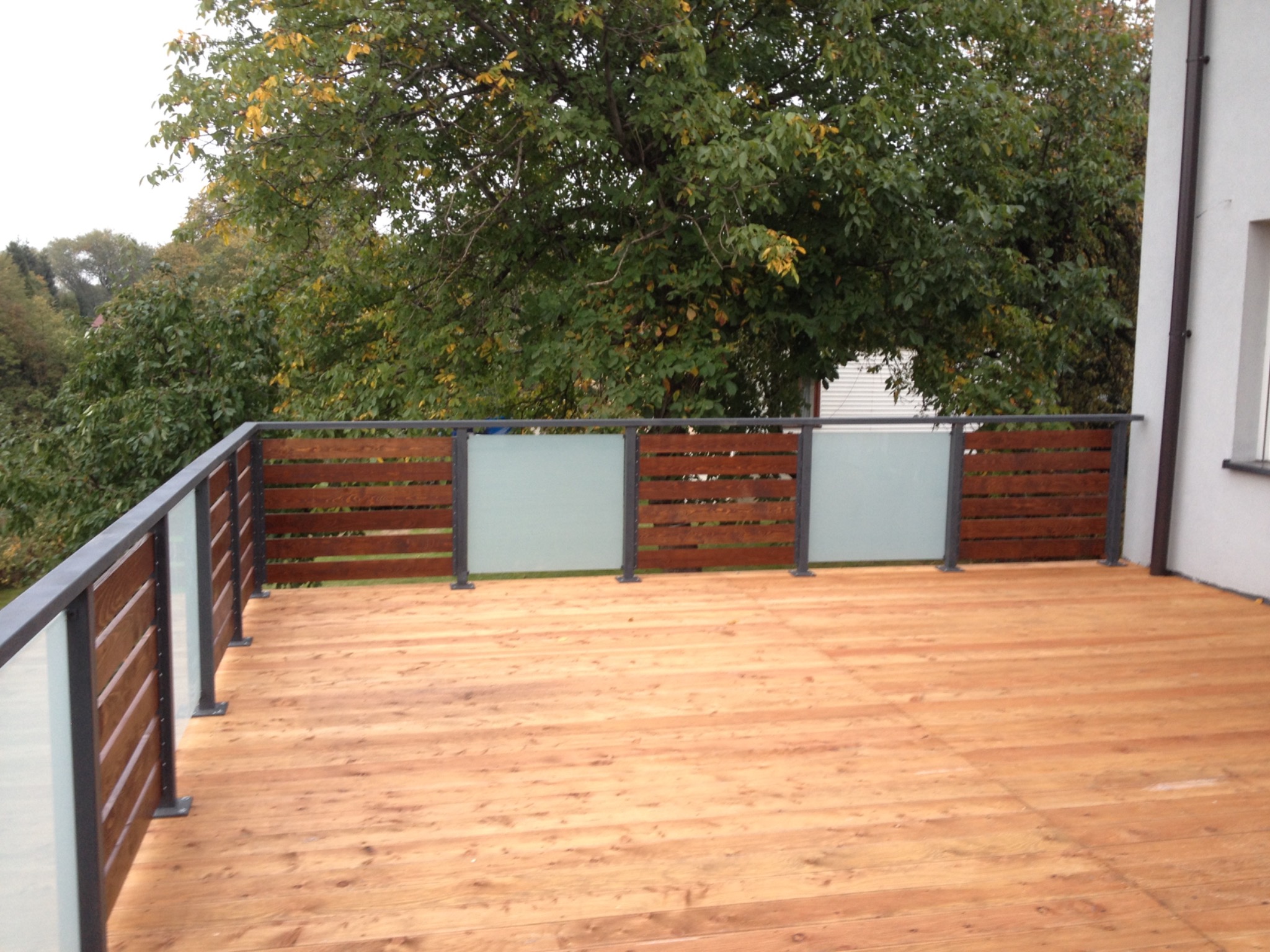 Nowoczesny balkon z drewnianą podłogą i balustradą z poziomych drewnianych paneli oraz matowego szkła, na tle drzew z żółknącymi liśćmi.