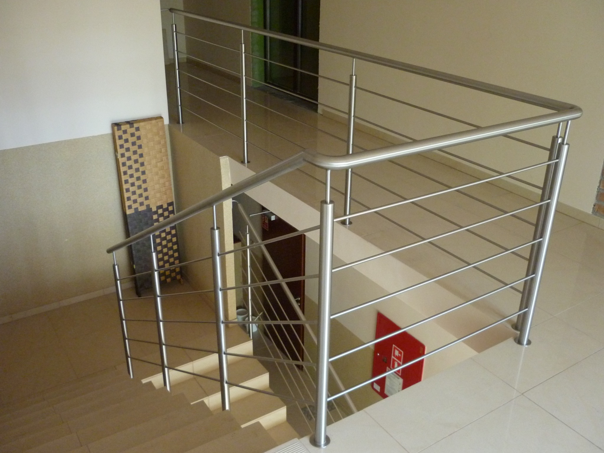 Nowoczesna balustrada ze stali nierdzewnej z poziomymi prętami, zamontowana przy schodach wewnątrz budynku, widoczna skrzynka przeciwpożarowa i dekoracyjny panel na ścianie.