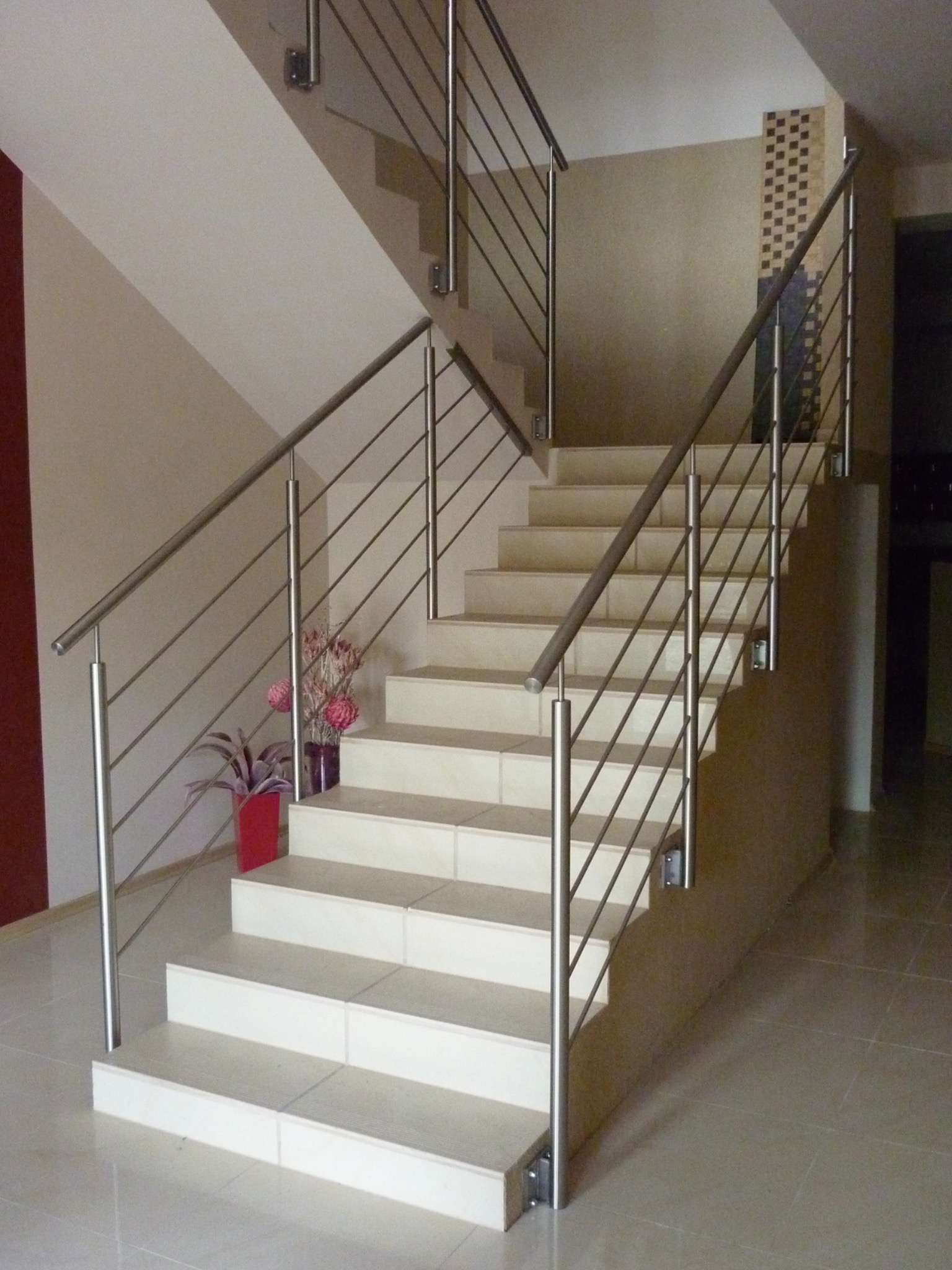 Schody wyłożone jasnymi płytkami z balustradą ze stali nierdzewnej, z doniczką z kwiatami na dole schodów, widok z dołu.