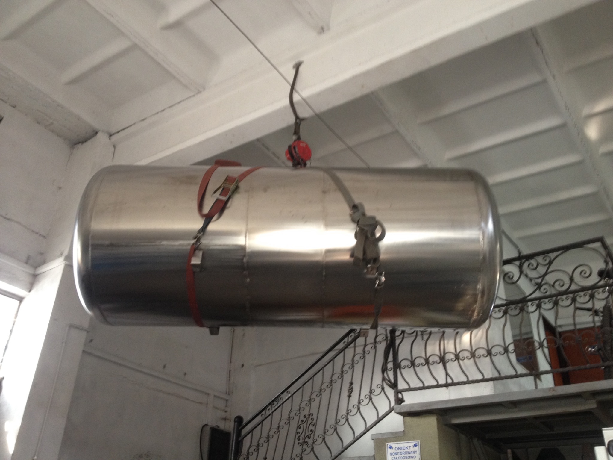 Błyszczący, metalowy zbiornik cylindryczny zawieszony na pasach wewnątrz budynku, widoczne schody z ozdobną, metalową balustradą w tle.