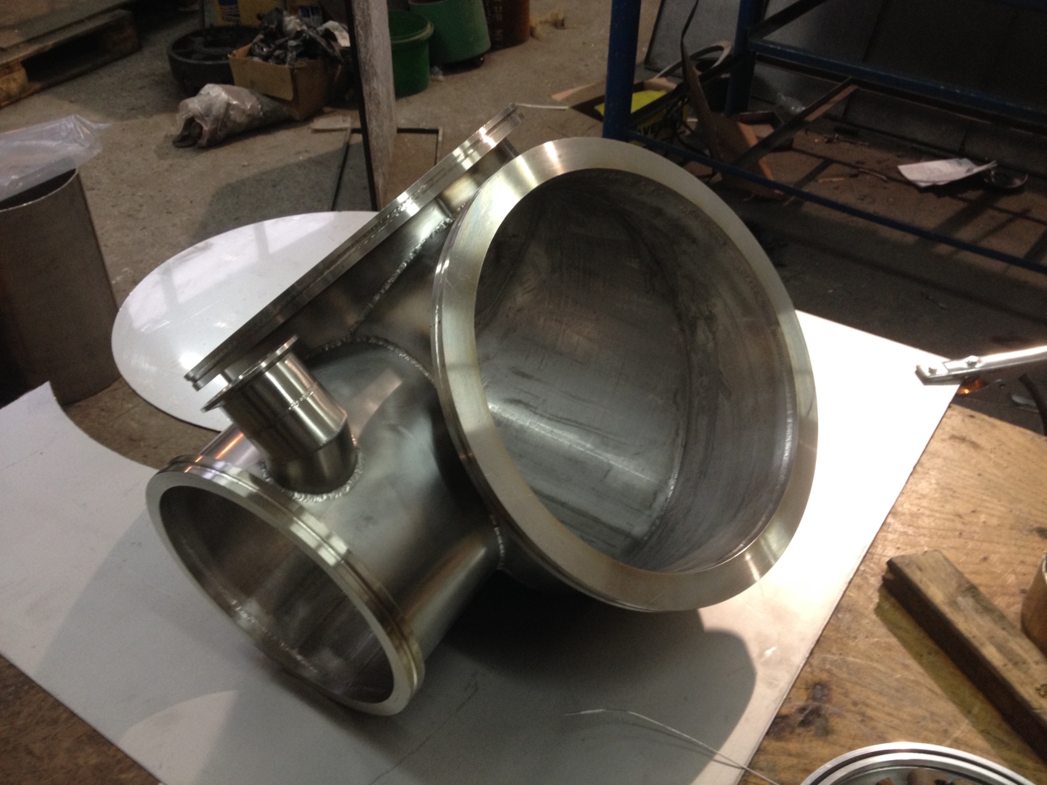 Precyzyjnie wykonany, spawany, nierdzewny trójnik rurowy z widocznymi śladami obróbki i spawania, leżący na stole warsztatowym.