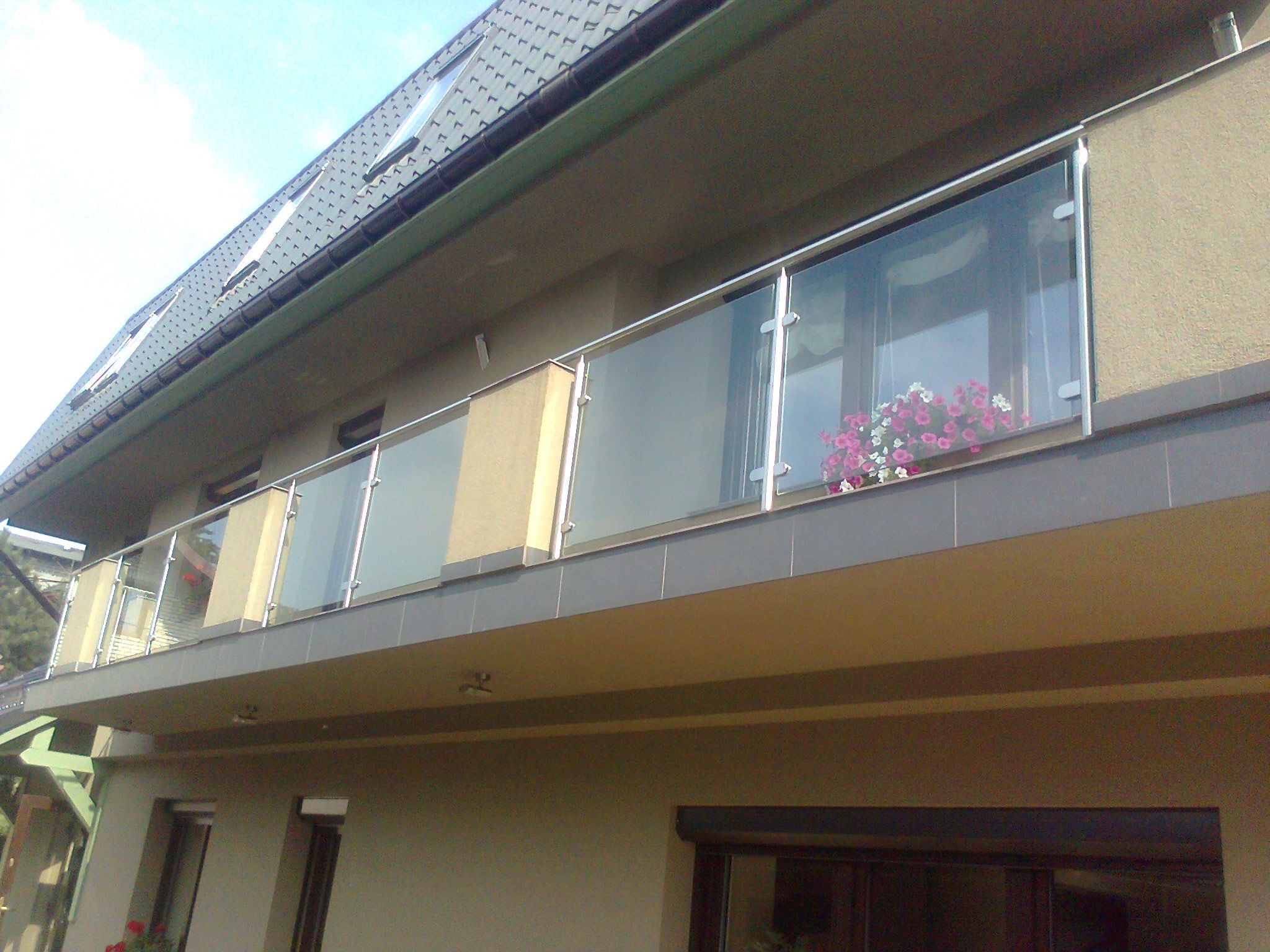 Szklana balustrada balkonowa z elementami ze stali nierdzewnej, widok z dołu na tle beżowej elewacji budynku z kwiatami w donicach.