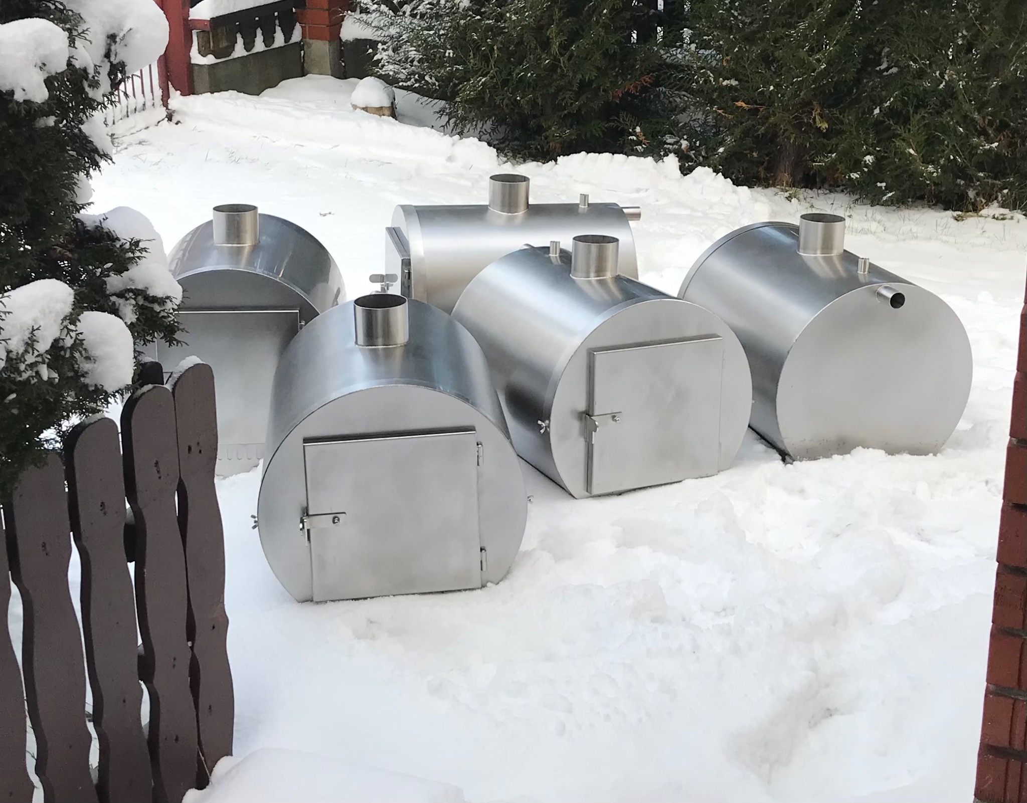 Pięć metalowych zbiorników z rurami i drzwiczkami, stojących na śniegu przed ogrodzeniem i krzewem.