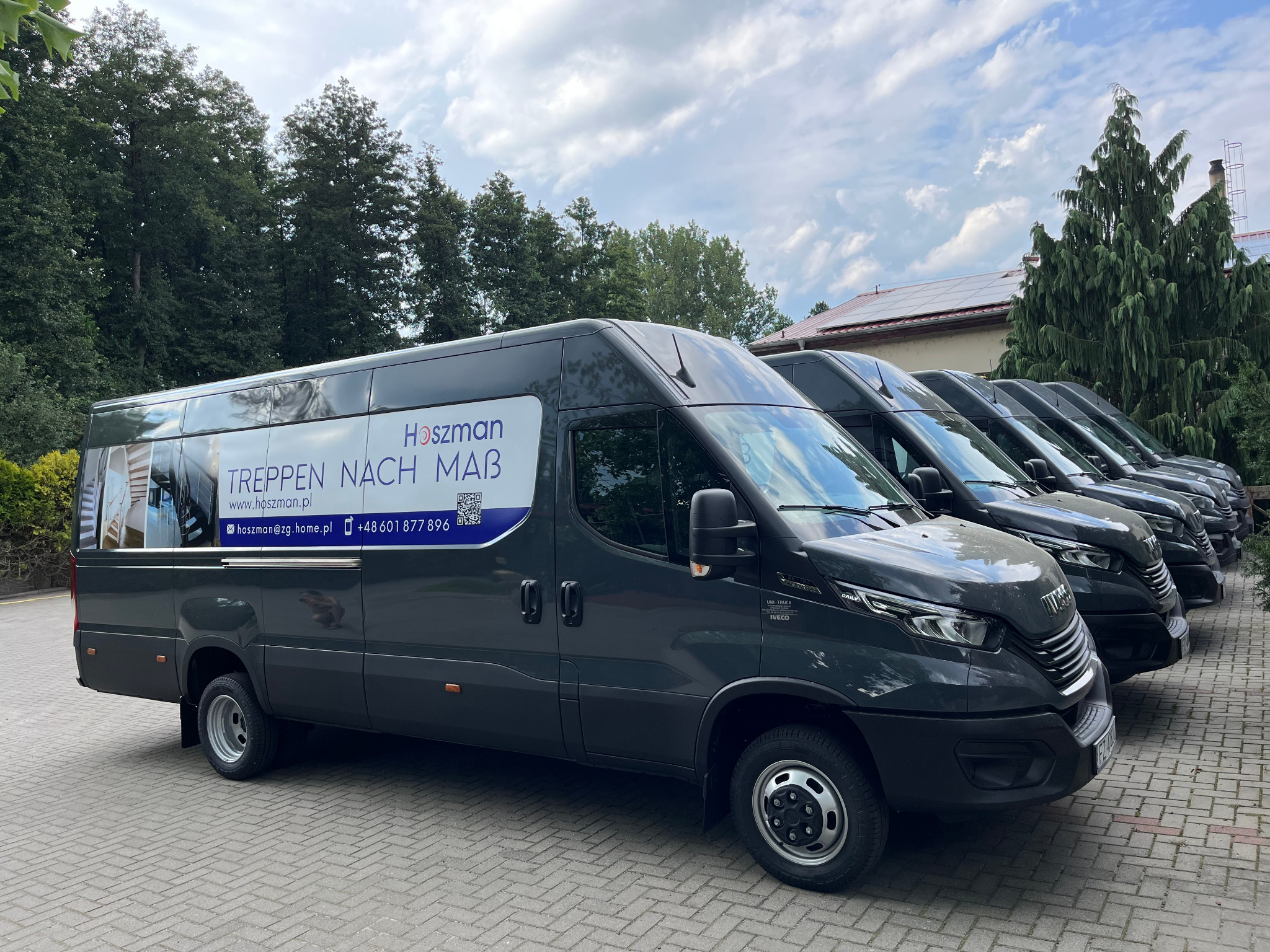 Flota szarych samochodów dostawczych firmy z reklamą schodów, zaparkowana na brukowanym placu, w tle drzewa i budynek z panelami słonecznymi.