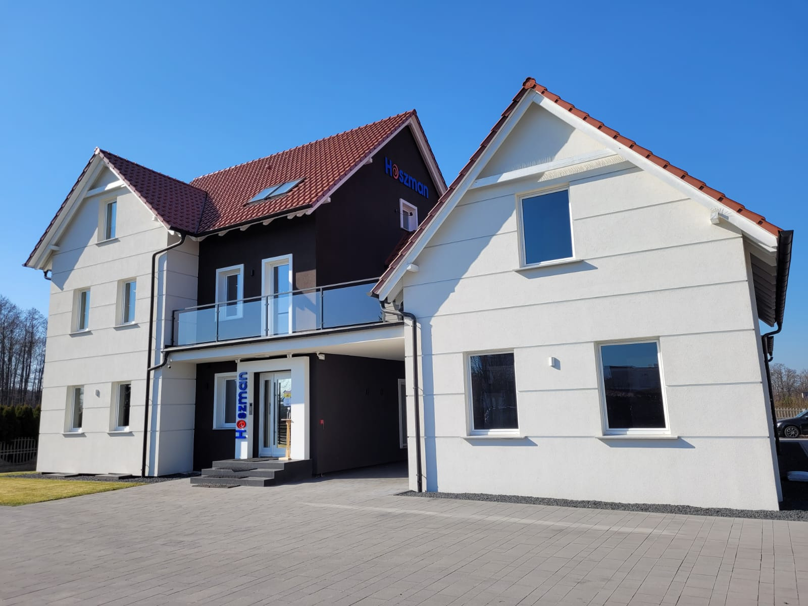 Nowoczesny budynek biurowy z dwoma segmentami o kontrastujących kolorach elewacji: białym i ciemnobrązowym, z czerwonym dachem i przeszklonym balkonem, na tle błękitnego nieba.