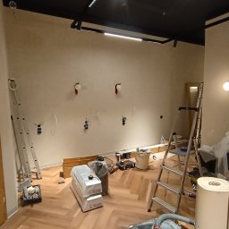 Adam Pacan - Wnętrze w trakcie remontu: parkiet ułożony w jodełkę, drabiny, odkurzacz przemysłowy, przygotowane kable elektryczne na ścianie, sufit pomalowany na czarno.