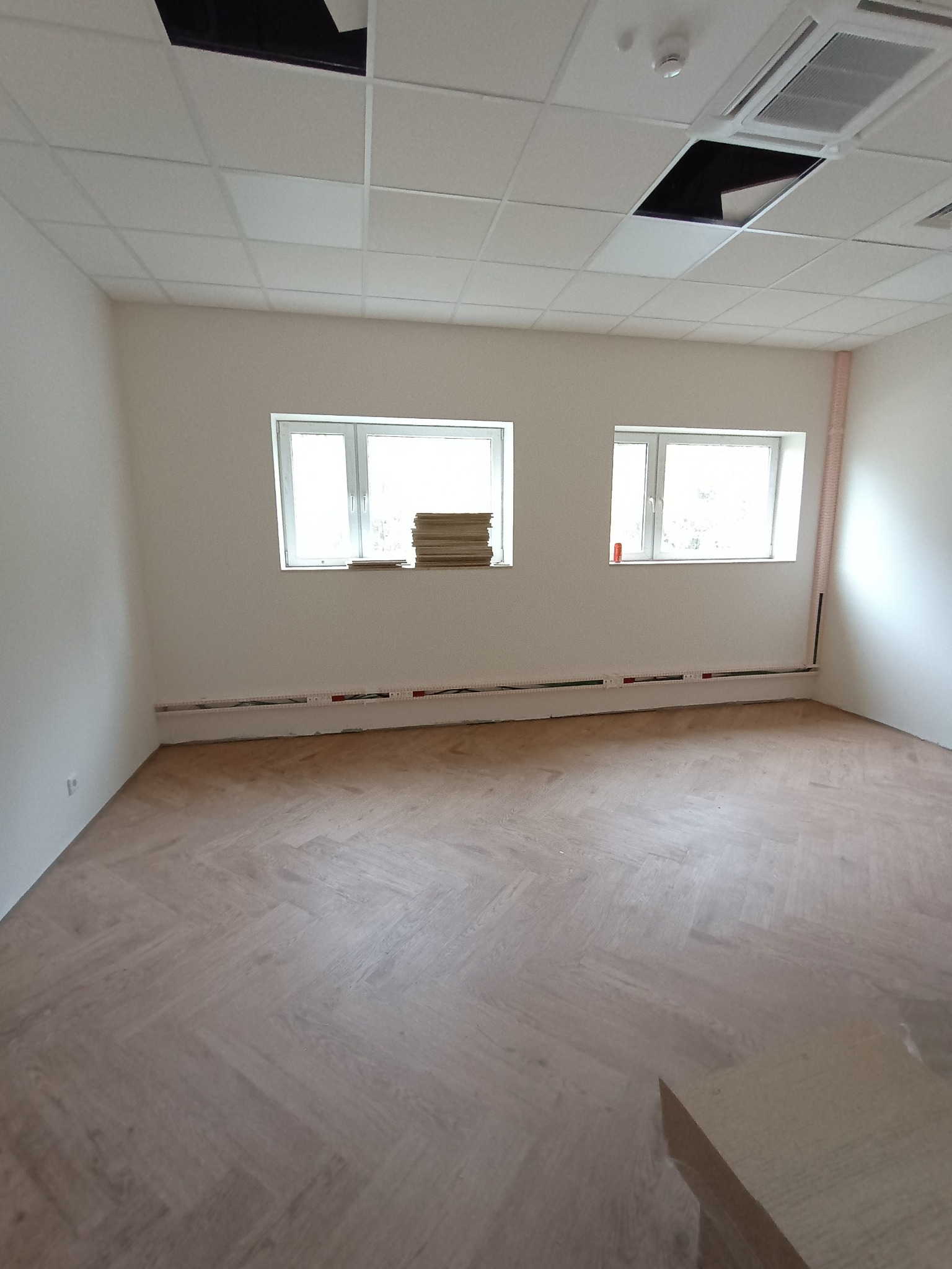 Puste biuro z nowo ułożoną podłogą w jodełkę, widoczne okna, grzejniki pod oknami i sufit kasetonowy z elementami instalacji.