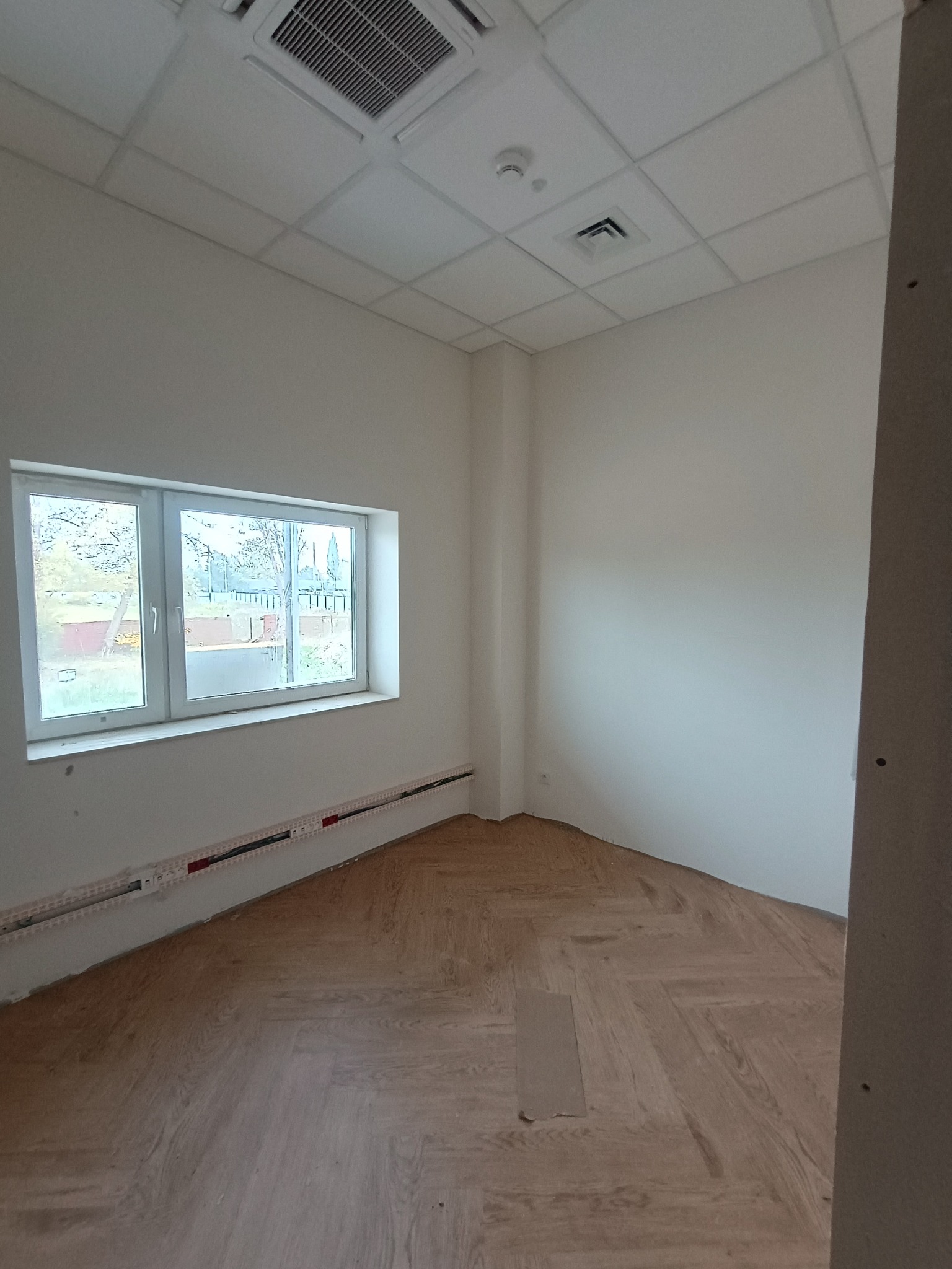 Puste pomieszczenie biurowe z parkietem ułożonym w jodełkę, białymi ścianami, oknem z widokiem na zewnątrz i widocznymi instalacjami elektrycznymi w listwach przypodłogowych.