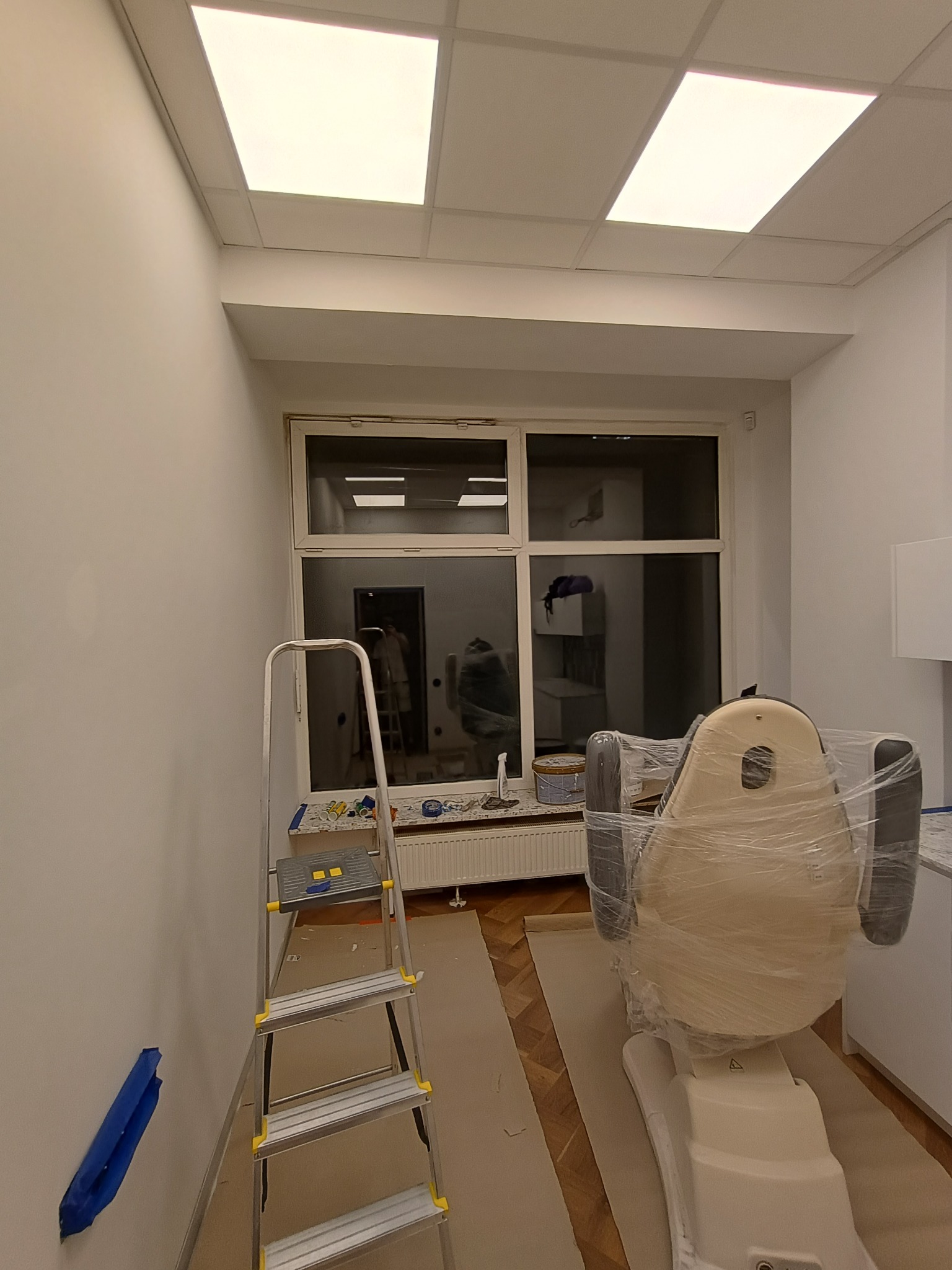 Wnętrze gabinetu z nowo pomalowanymi ścianami na biało, drabina aluminiowa przy ścianie, zabezpieczony folią fotel zabiegowy, okno z widokiem na inne pomieszczenie.