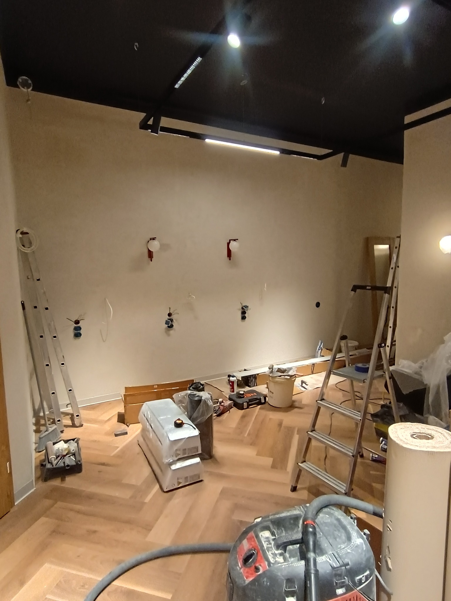 Wnętrze w trakcie remontu: parkiet ułożony w jodełkę, drabiny, odkurzacz przemysłowy, przygotowane kable elektryczne na ścianie, sufit pomalowany na czarno.