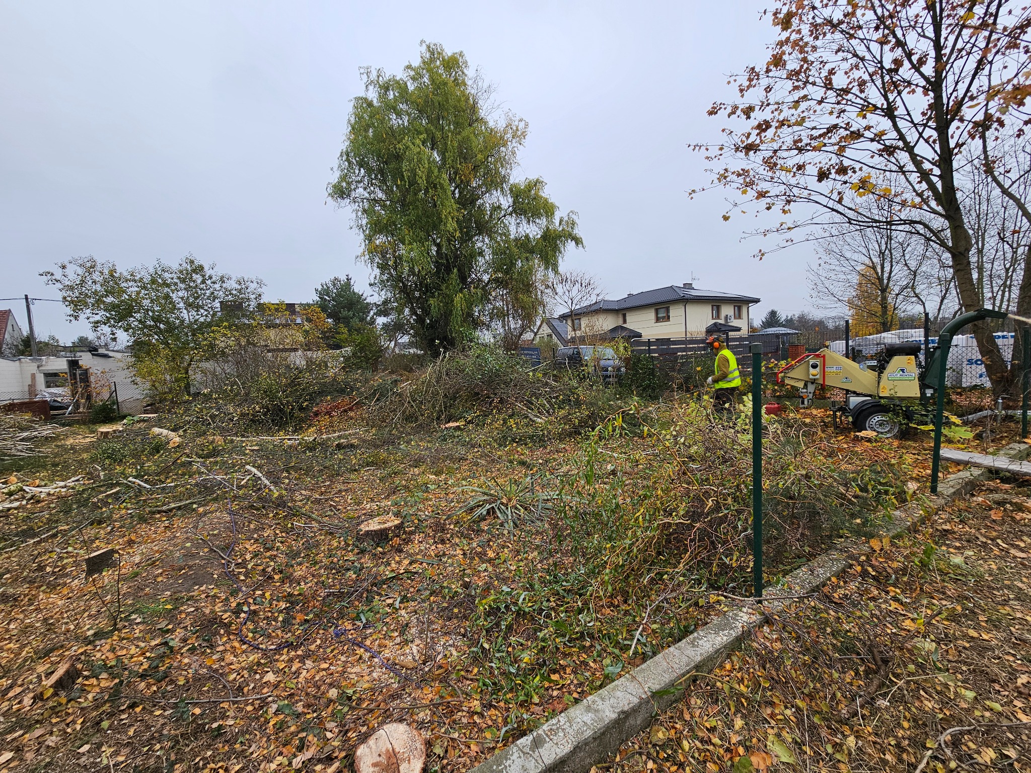 Teren po wycince drzew i krzewów, widoczny pracownik w odblaskowej kamizelce i rębarka do gałęzi, liście na ziemi, w tle zabudowa jednorodzinna.