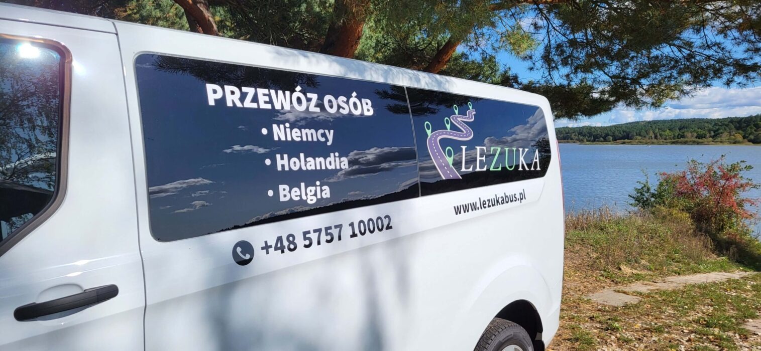 Biały bus z naklejką informującą o transporcie do Niemiec, Holandii i Belgii, zaparkowany nad jeziorem w słoneczny dzień.