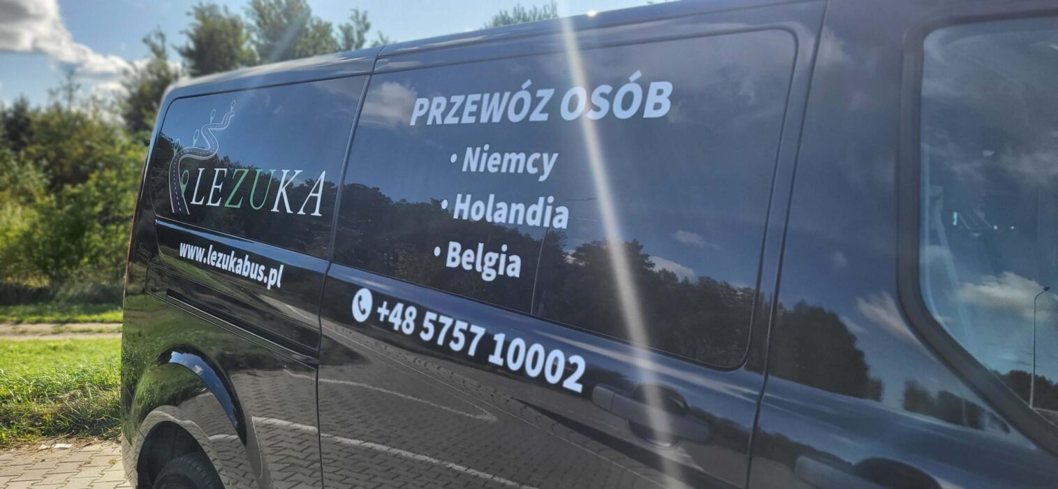 Ciemny bus z naklejonym logo firmy przewozowej 'LEZUKA' i informacją o przewozach do Niemiec, Holandii i Belgii oraz numerem telefonu, zaparkowany na parkingu w słoneczny dzień.