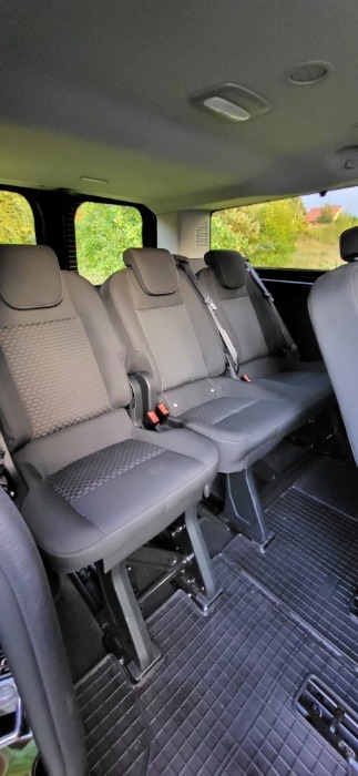 Wnętrze busa z trzema rzędami szarych foteli z regulowanymi zagłówkami i pasami bezpieczeństwa, widok z perspektywy tylnych siedzeń, gumowa podłoga z wzorem kratki.