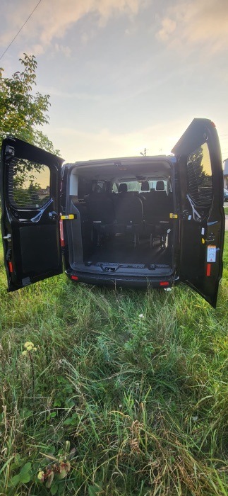 Czarny bus z otwartymi tylnymi drzwiami, widok na wnętrze z fotelami pasażerskimi, stojący na trawiastym terenie.