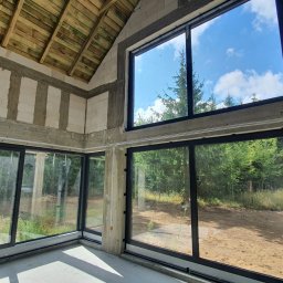 PRO-HOUSE SPÓŁKA Z OGRANICZONĄ ODPOWIEDZIALNOŚCIĄ - Surowy stan wnętrza z dużymi, czarnymi oknami aluminiowymi, widok na las i niebo, widoczne przewody elektryczne na ścianach z betonu komórkowego.