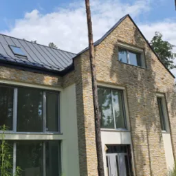 Nowoczesny dom jednorodzinny w Warszawie z elewacją z kamienia i dużymi oknami, widok z zewnątrz w słoneczny dzień.