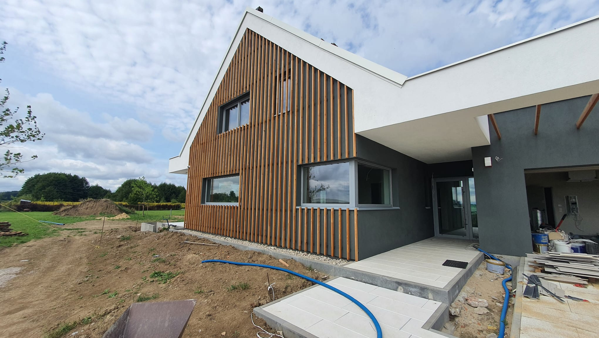 Nowoczesny dom z elewacją z drewnianych listew, szarymi oknami i częściowo wykończonym terenem wokół, widoczny niebieski wąż ogrodowy.