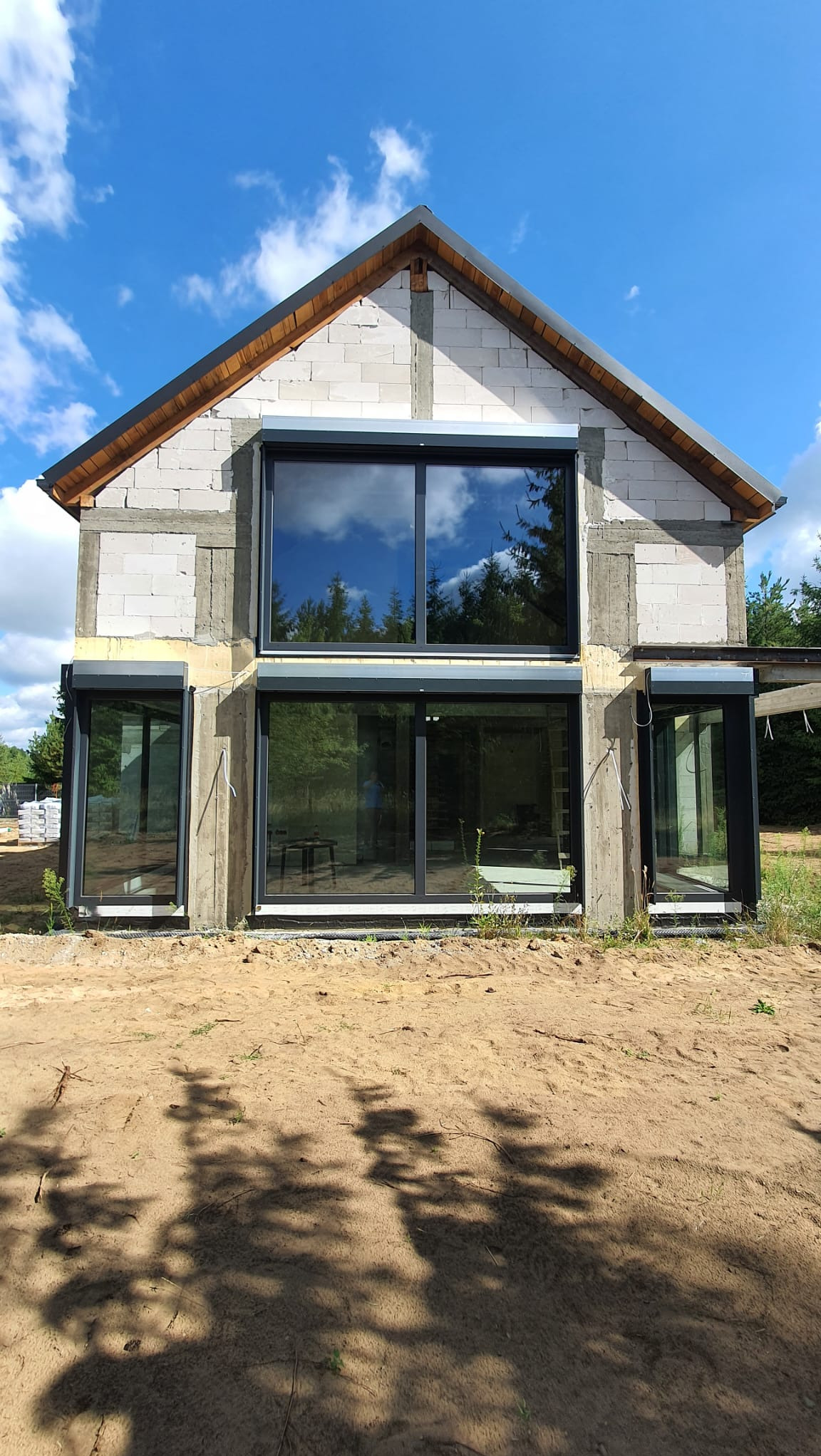 Surowy budynek w trakcie budowy z zamontowanymi dużymi oknami z czarnymi ramami i częściowo opuszczonymi roletami zewnętrznymi w kolorze szarym, odbijające się chmury na szybach.