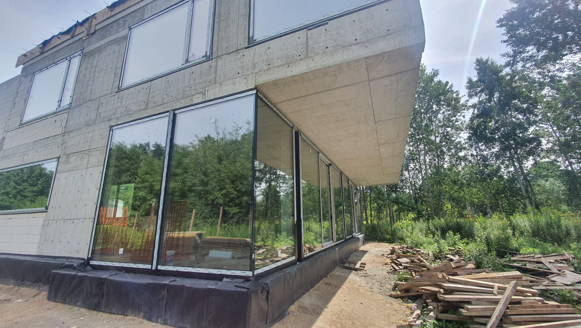Surowy betonowy dom z dużymi oknami aluminiowymi, widok z zewnątrz na las, teren budowy z deskami.