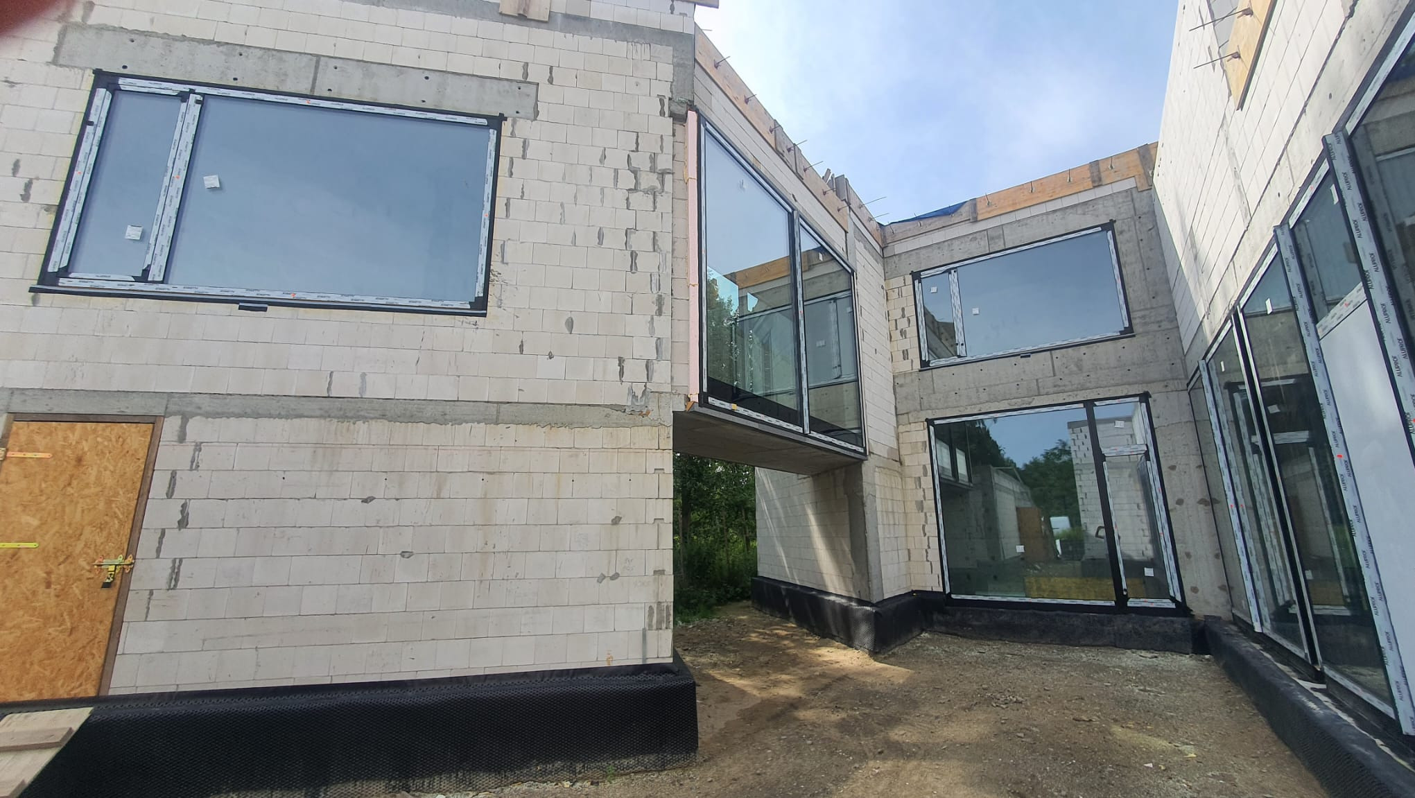 Montaż okien PCV w surowym stanie budynku z białego bloczka, widoczne zabezpieczenie fundamentów folią hydroizolacyjną.