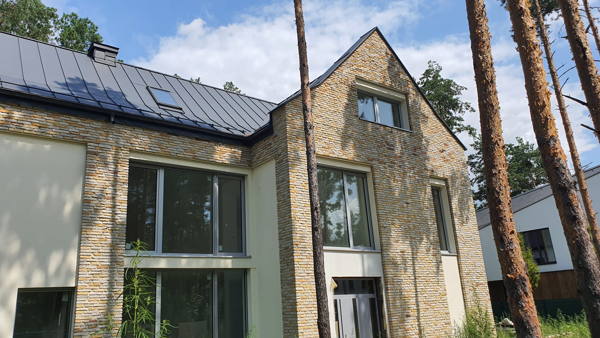 Nowoczesny dom jednorodzinny w Warszawie z elewacją z kamienia i dużymi oknami, widok z zewnątrz w słoneczny dzień.