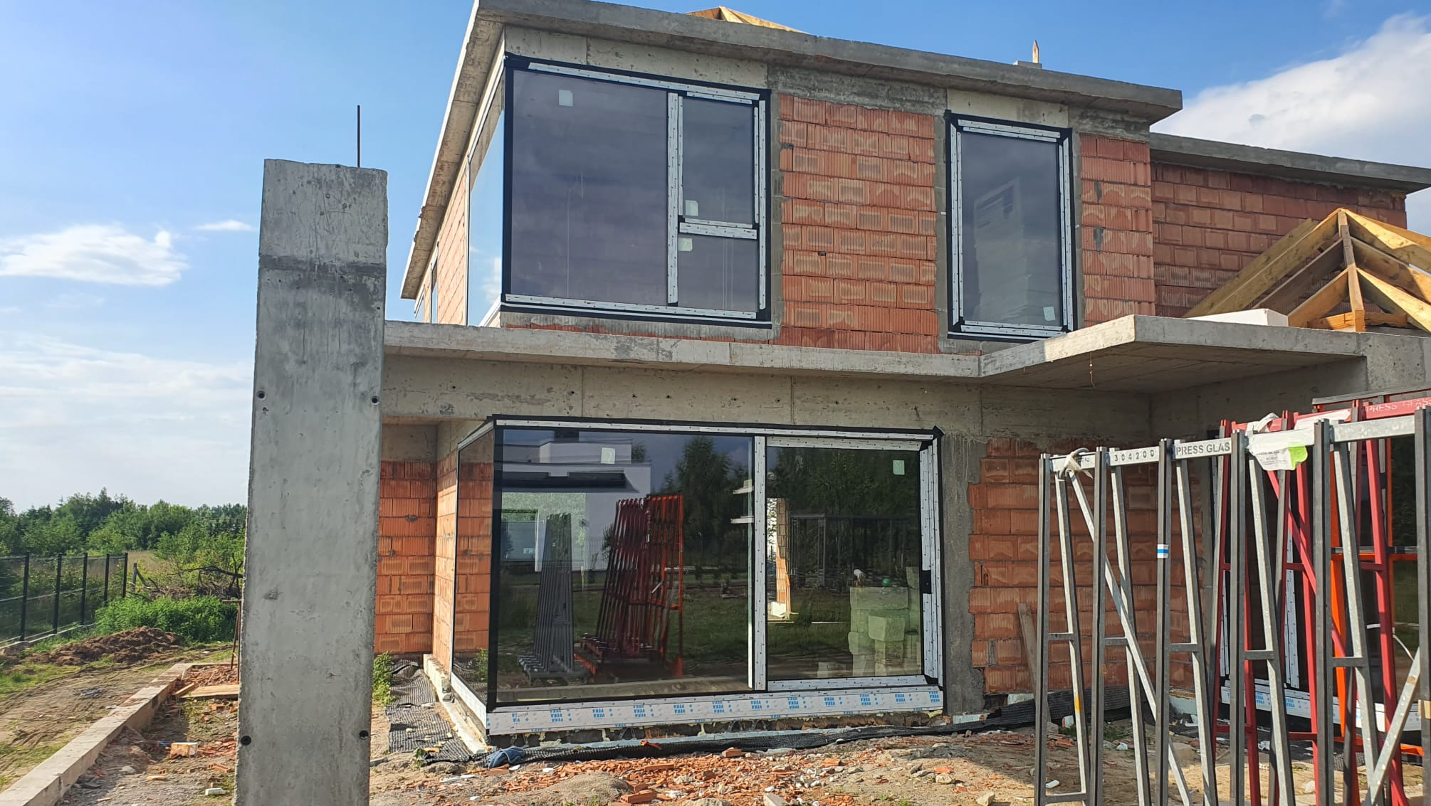 Surowy stan budowy domu jednorodzinnego z zamontowanymi oknami z białymi ramami, widoczne cegły, betonowe elementy konstrukcyjne i stojaki z szybami.