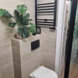 Jakub Wiśniewski Usługi Remontowo-Budowlane - Jasna łazienka z beżowymi płytkami, wiszącą toaletą i czarnym grzejnikiem drabinkowym. Dekoracyjny akcent stanowi zielona roślina w doniczce.