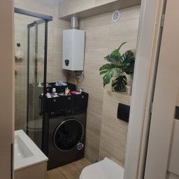 Jakub Wiśniewski Usługi Remontowo-Budowlane - Wykończona łazienka w beżowych odcieniach, z kabiną prysznicową, pralką i rośliną doniczkową,  grzejnik nad pralką. Minimalistyczny wystrój.