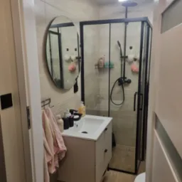 Nowoczesna łazienka z kabiną prysznicową typu walk-in, okrągłym lustrem i beżowymi płytkami. Widoczna umywalka nablatowa z szafką i drewnopodobna podłoga.