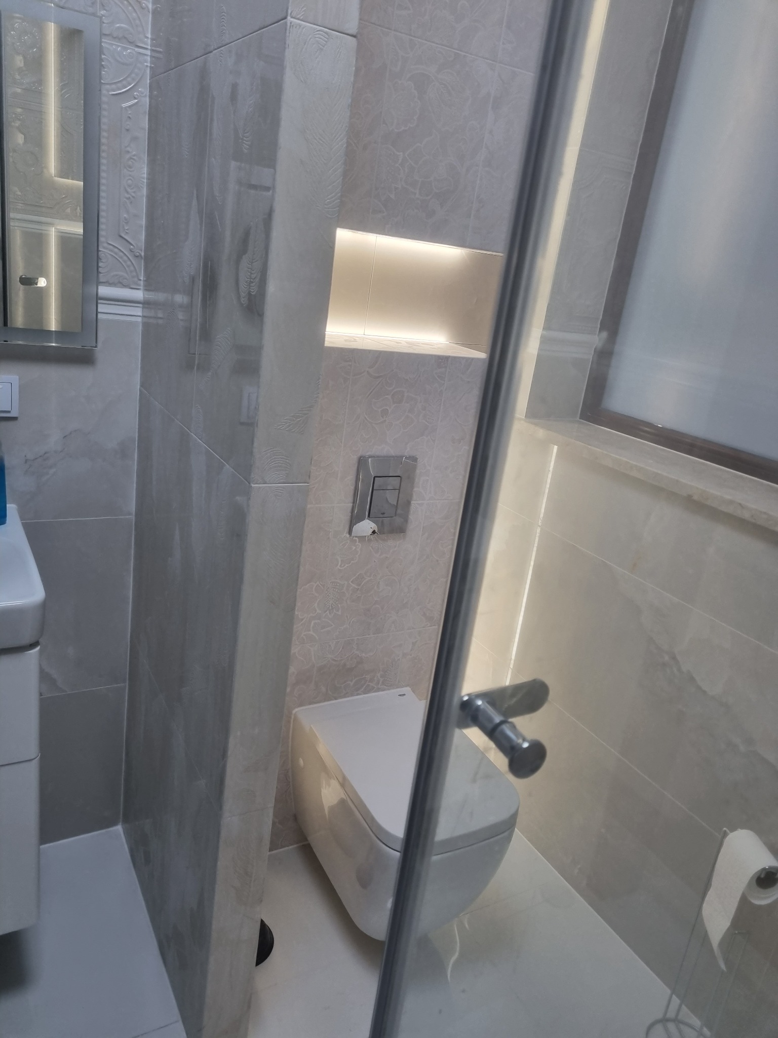 Elegancka łazienka w odcieniach szarości, z podświetlaną wnęką nad toaletą i kabiną prysznicową ze szklanymi drzwiami. Widoczna umywalka z lustrem.