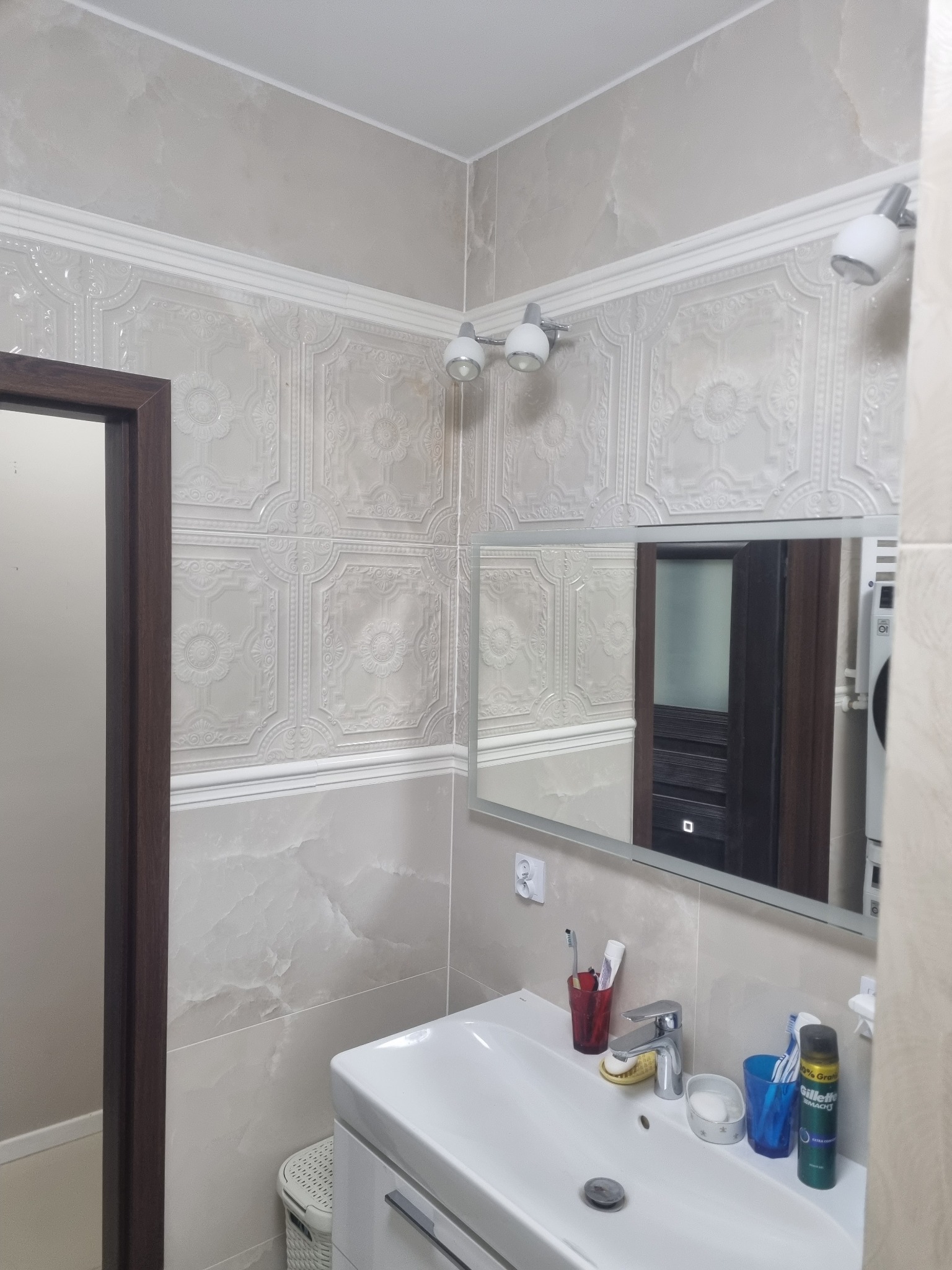 Jasna łazienka z umywalką i lustrem. Ściany wyłożone płytkami z delikatnym wzorem, oświetlenie punktowe. Widoczne akcesoria łazienkowe.