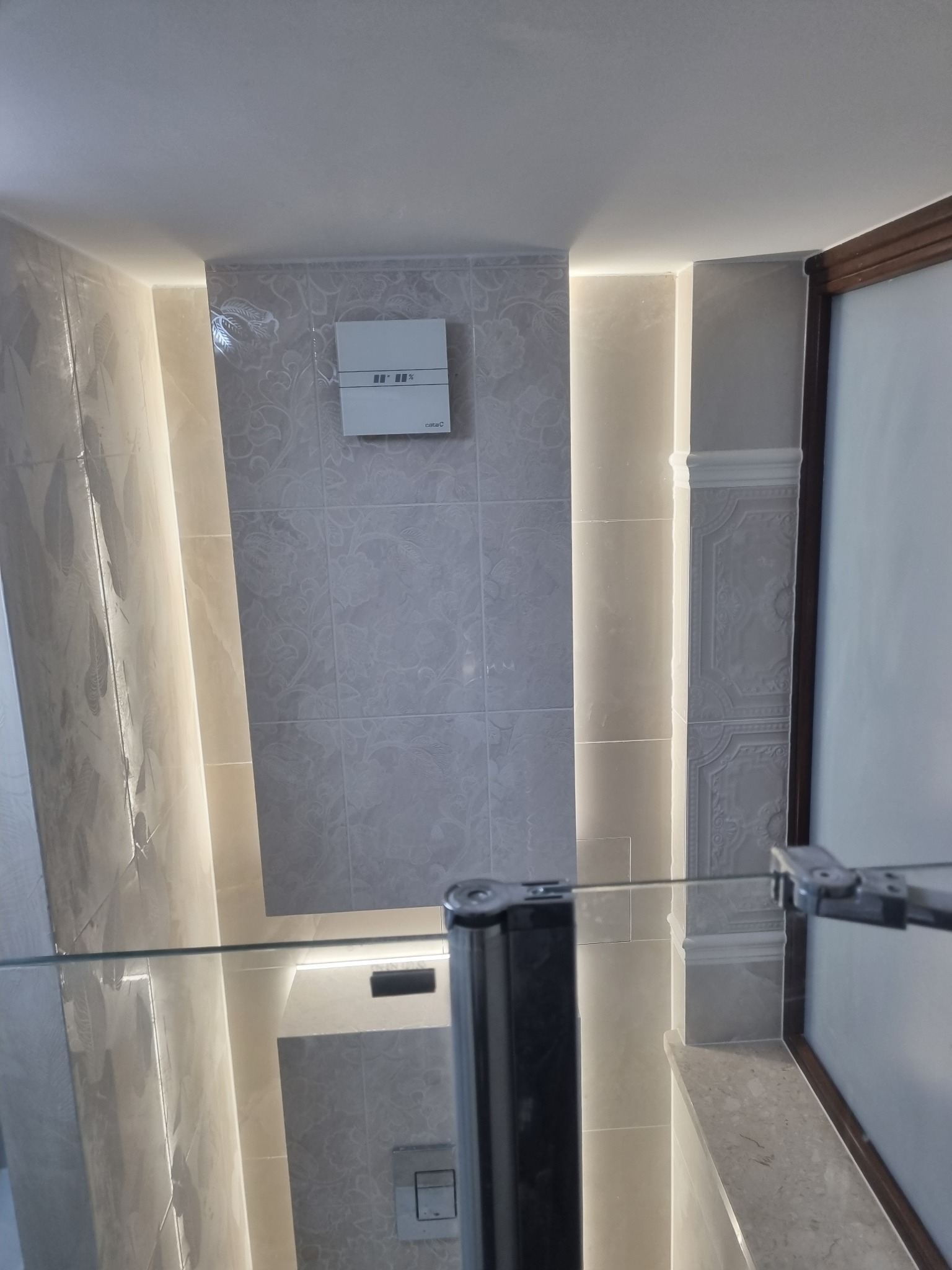 Wykończona łazienka z szarymi płytkami w kwiatowy wzór, podświetleniem LED w pionowych wnękach i szklanym panelem. Widoczny wentylator na ścianie.