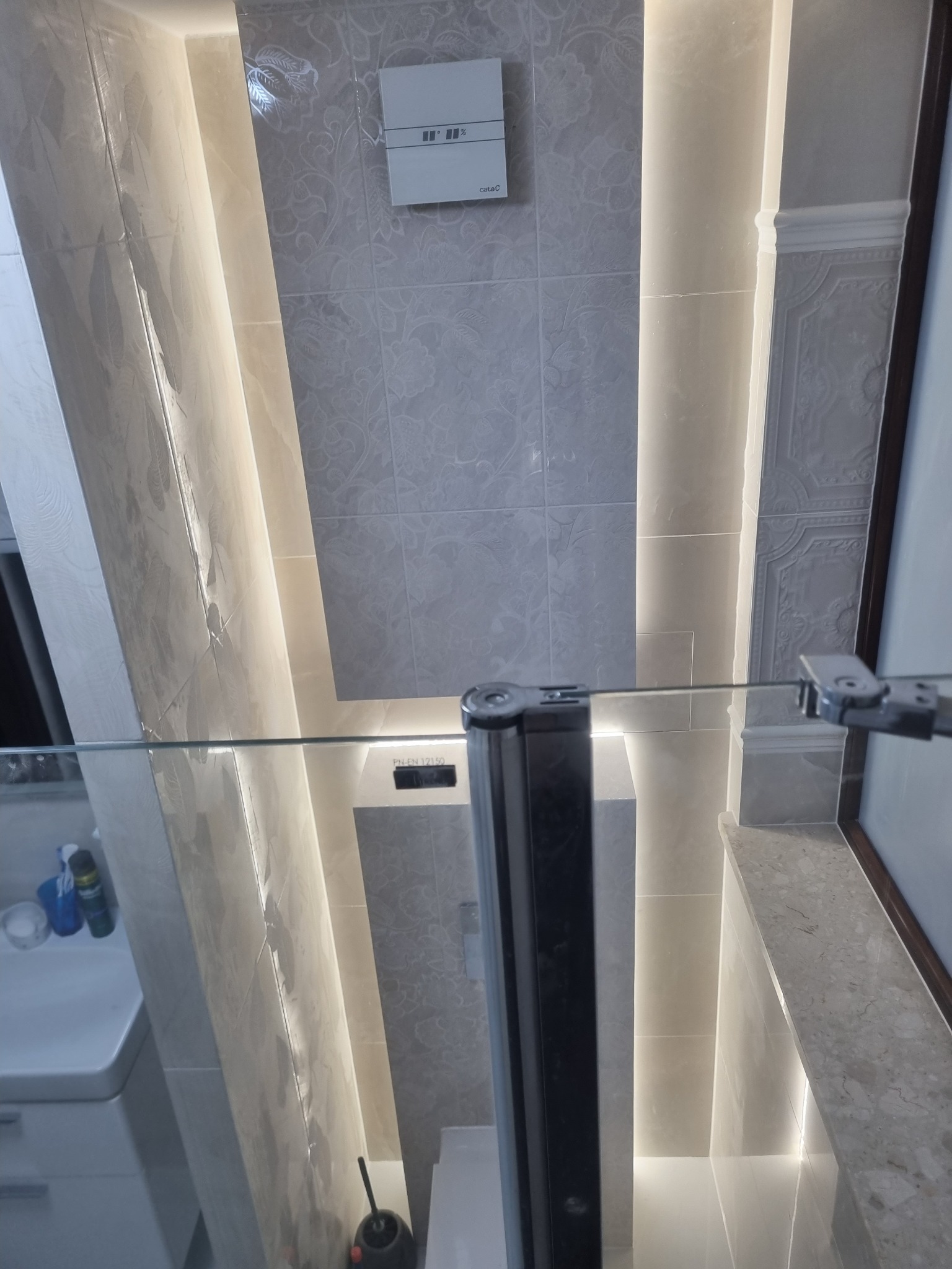 Elegancka łazienka z podświetlanymi płytkami w odcieniach szarości, kabina prysznicowa ze szklanymi drzwiami, widoczna umywalka i toaleta. Nowoczesny design.