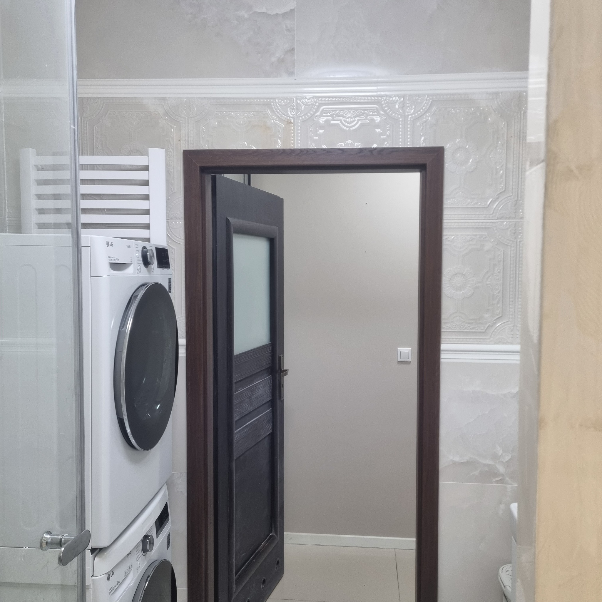 Wykończona łazienka z pralką LG, grzejnikiem i otwartymi drzwiami. Płytki imitujące marmur i dekoracyjne panele ścienne. Neutralna kolorystyka.