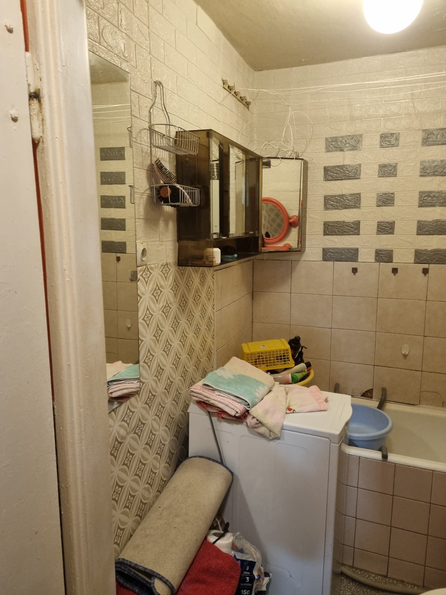 Niefotogeniczna łazienka z pralką, wiszącą szafką z lustrem i wzorzystymi płytkami. Widoczne akcesoria łazienkowe i tekstylia. Niewykorzystany potencjał aranżacyjny.