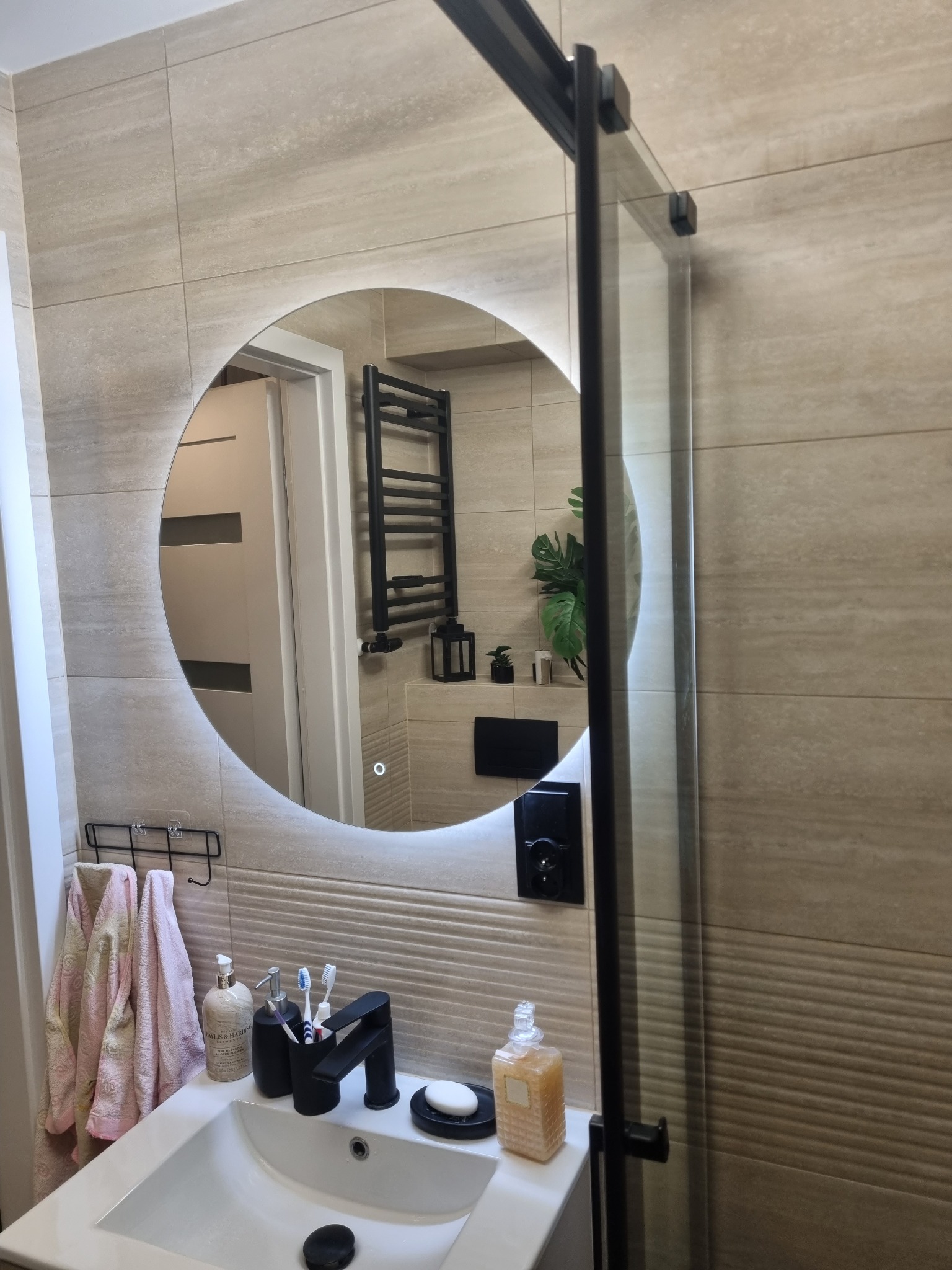 Beżowa łazienka z okrągłym lustrem LED, czarną armaturą i kabiną prysznicową. Widoczne płytki w odcieniach beżu i wyposażenie łazienkowe.