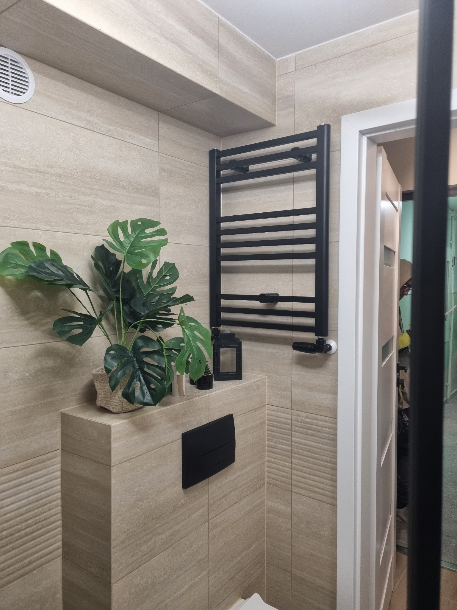 Beżowa łazienka z rośliną monstera obok czarnego grzejnika drabinkowego i białych drzwi z czarną ramą, wykończona płytkami imitującymi drewno.