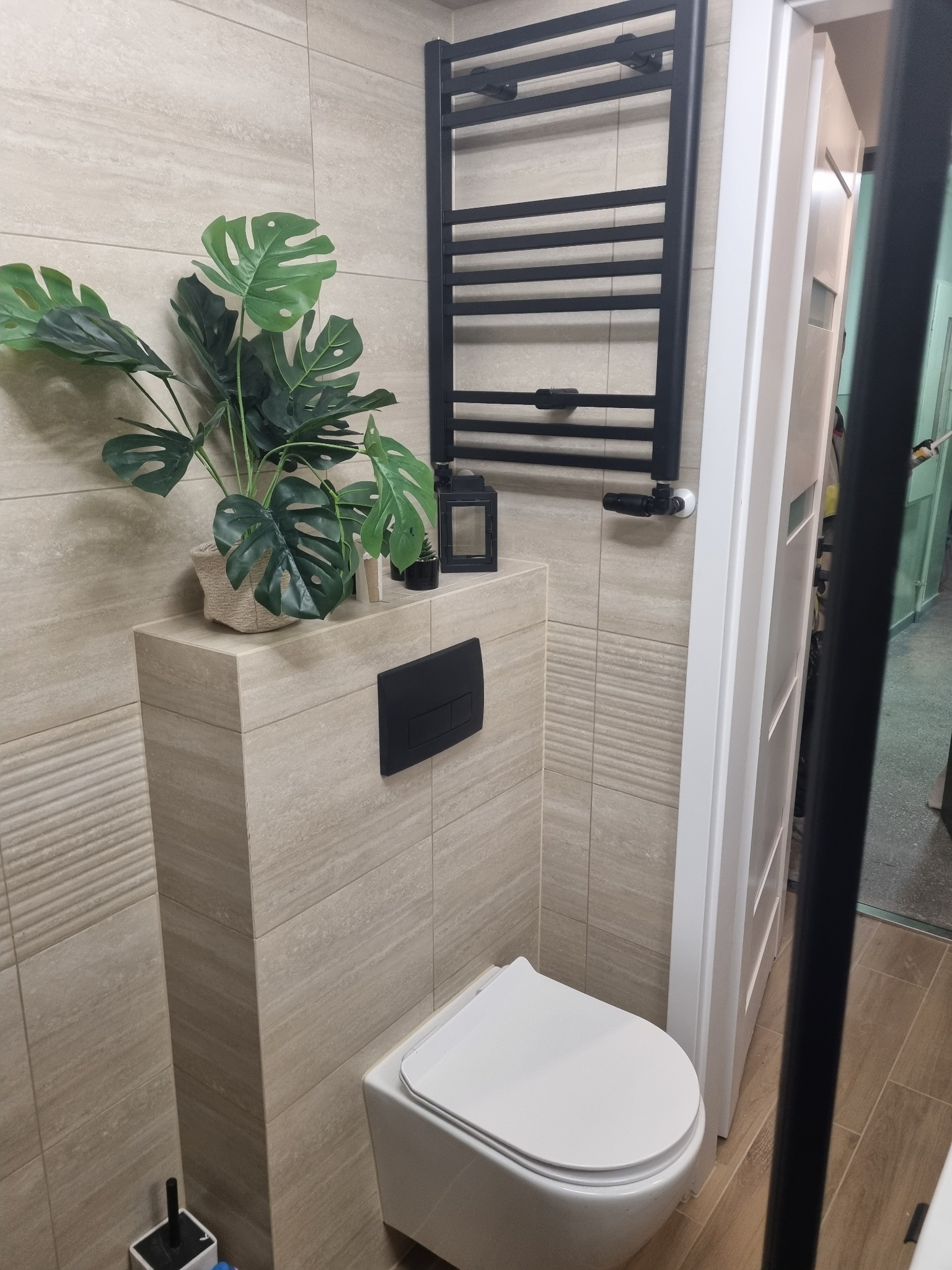 Jasna łazienka z beżowymi płytkami, wiszącą toaletą i czarnym grzejnikiem drabinkowym. Dekoracyjny akcent stanowi zielona roślina w doniczce.