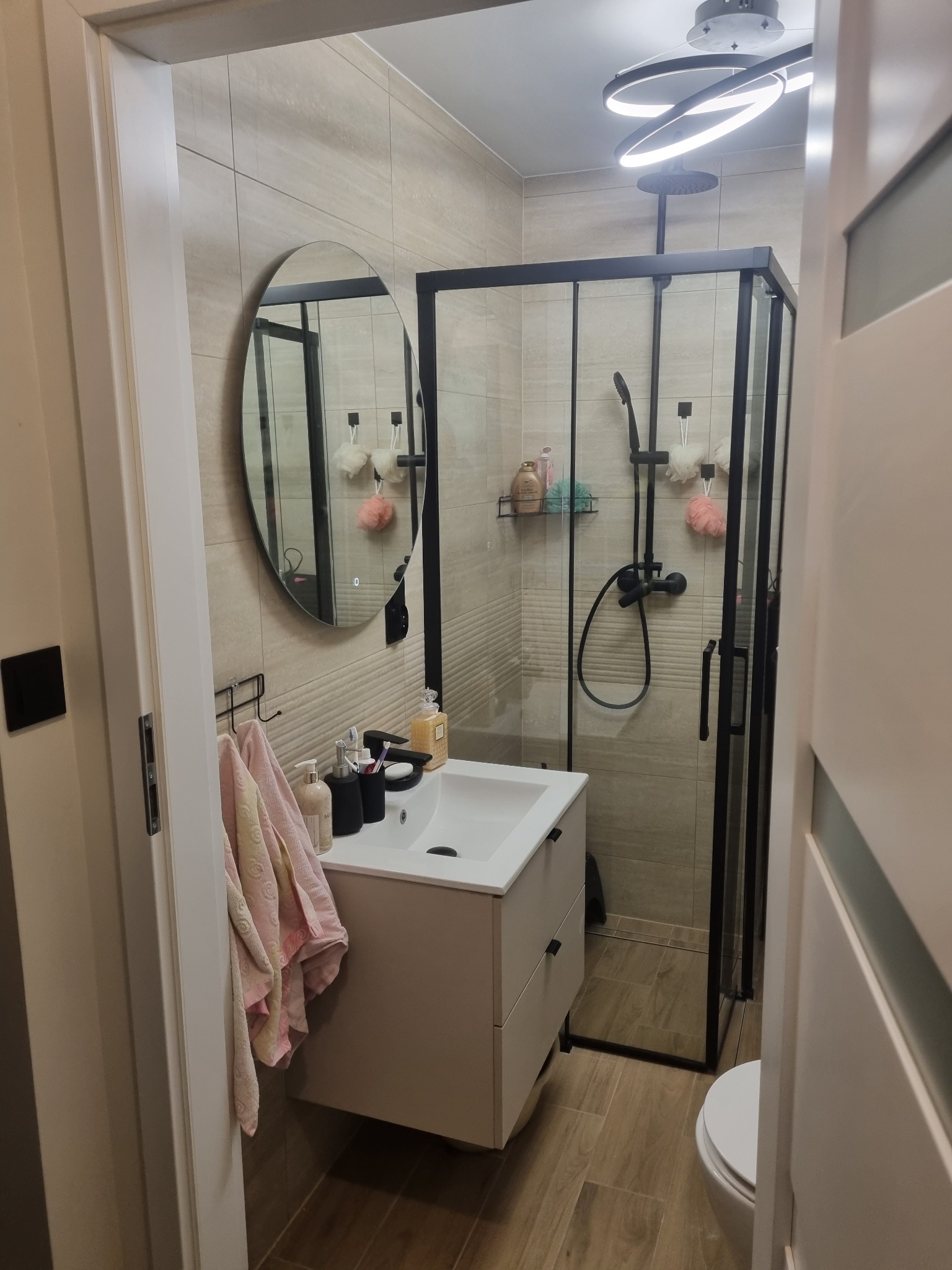 Nowoczesna łazienka z kabiną prysznicową typu walk-in, okrągłym lustrem i beżowymi płytkami. Widoczna umywalka nablatowa z szafką i drewnopodobna podłoga.