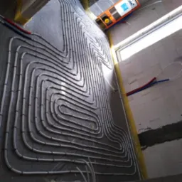 Układ rur ogrzewania podłogowego w trakcie montażu na przygotowanej powierzchni, widoczne mocowania i fragment instalacji sanitarnej.