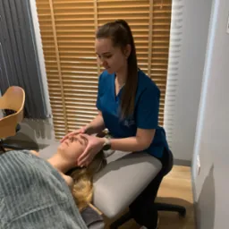 Kobieta w turkusowym uniformie delikatnie masuje czoło klientki leżącej na szarym stole do masażu, przykrytej ręcznikiem w paski; w tle drewniane żaluzje.