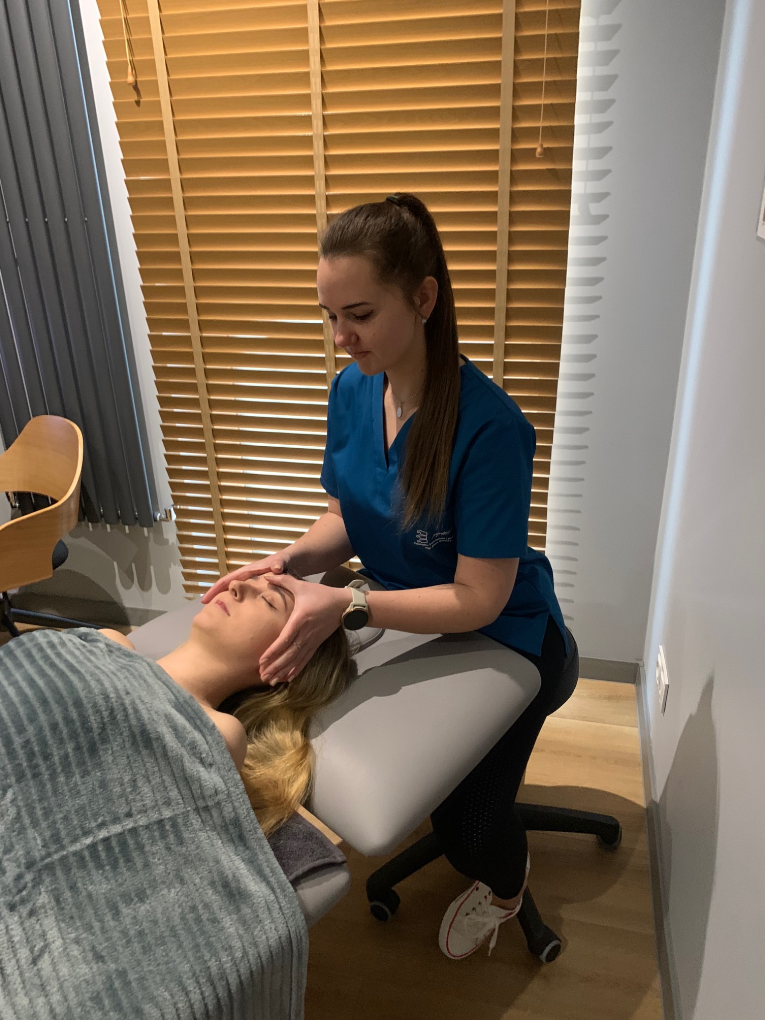 Kobieta w turkusowym uniformie delikatnie masuje czoło klientki leżącej na szarym stole do masażu, przykrytej ręcznikiem w paski; w tle drewniane żaluzje.