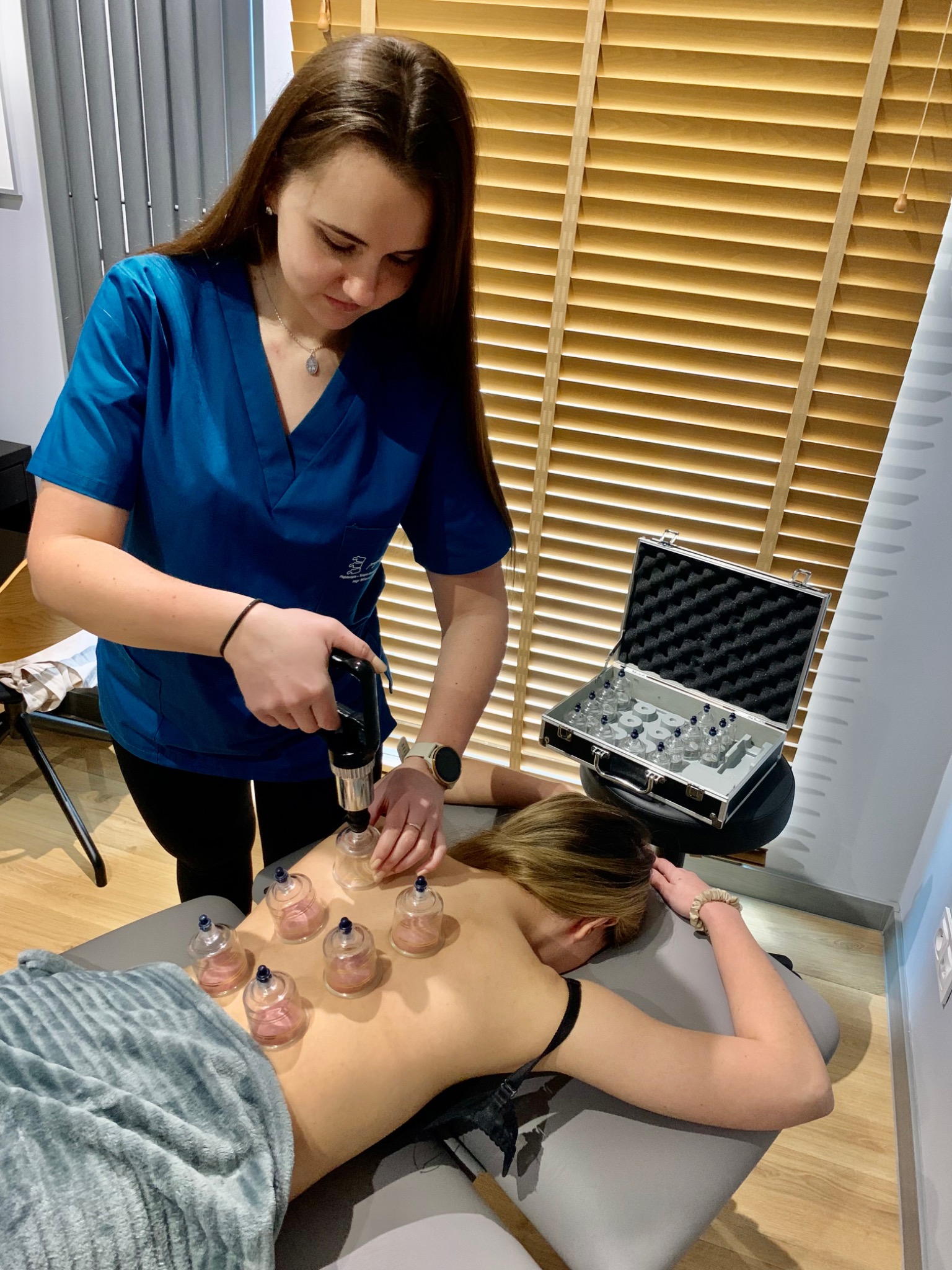 Kobieta w niebieskim uniformie aplikuje bańki próżniowe na plecy pacjentki leżącej na szarym stole do masażu, obok otwarty metalowy futerał z zapasowymi bańkami, w tle drewniane żaluzje.