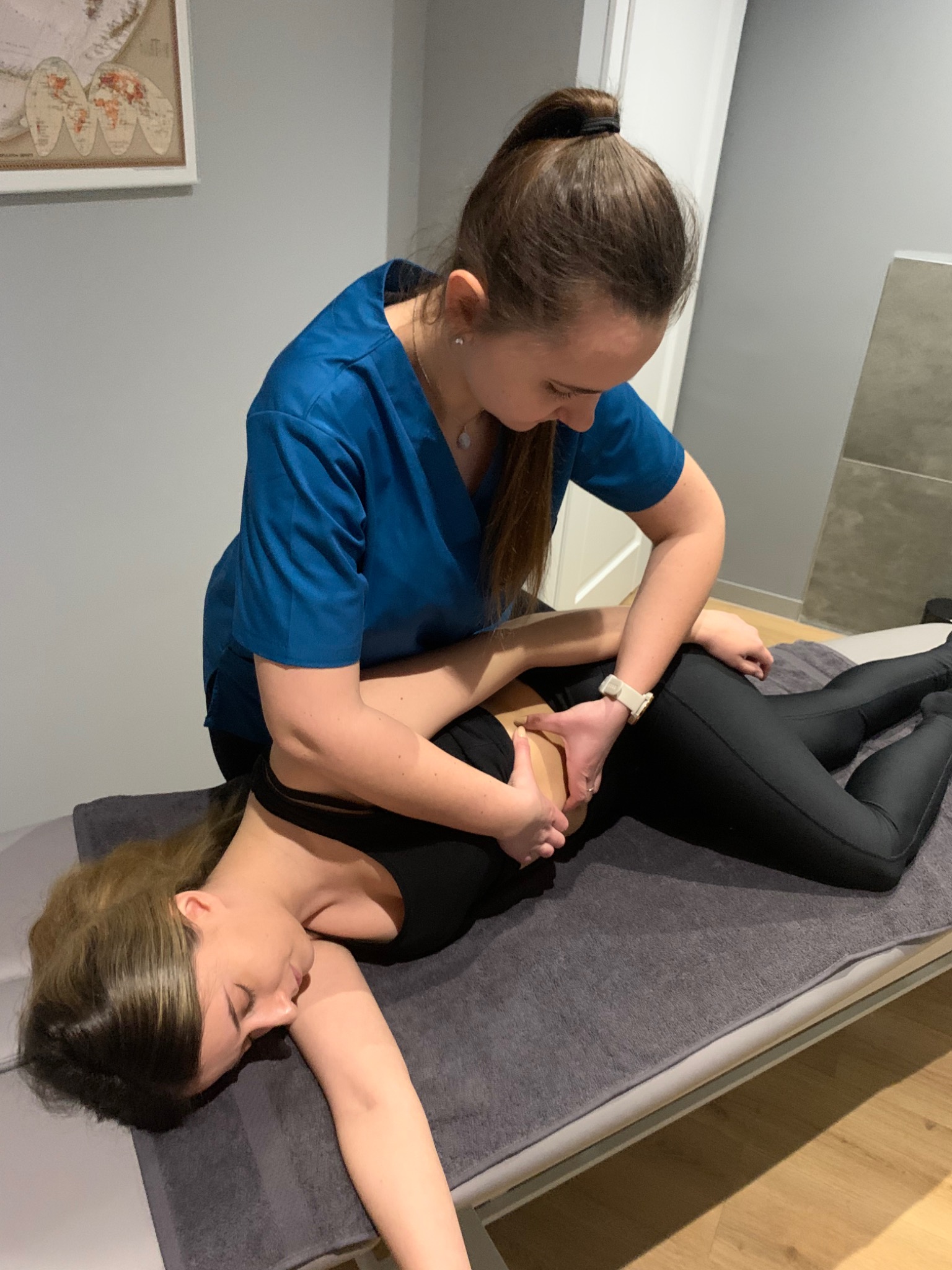 Kobieta w niebieskiej bluzie uciska dłonią dolną część pleców pacjentki leżącej na szarym łóżku do masażu, w tle widoczna mapa świata na ścianie.