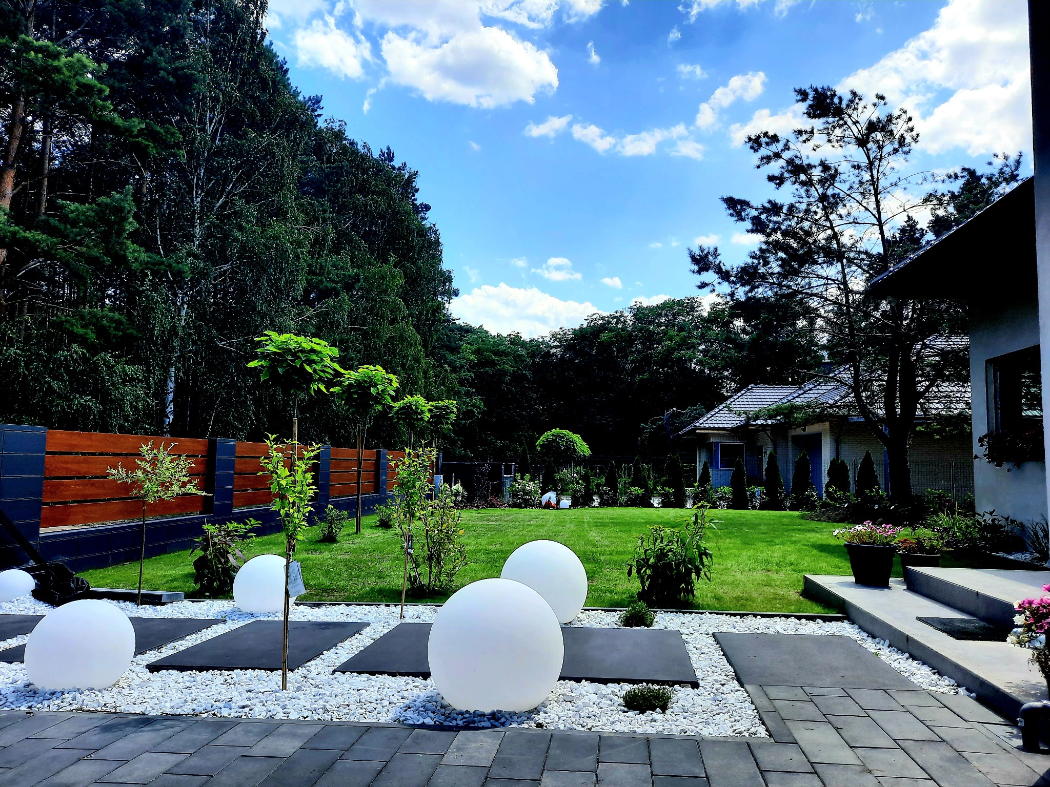 Starannie zaprojektowany ogród z równo przystrzyżonym trawnikiem, dekoracyjnymi kulami świetlnymi, ścieżkami z białego żwiru i płyt oraz młodymi drzewkami wzdłuż drewnianego ogrodzenia, na tle domu...