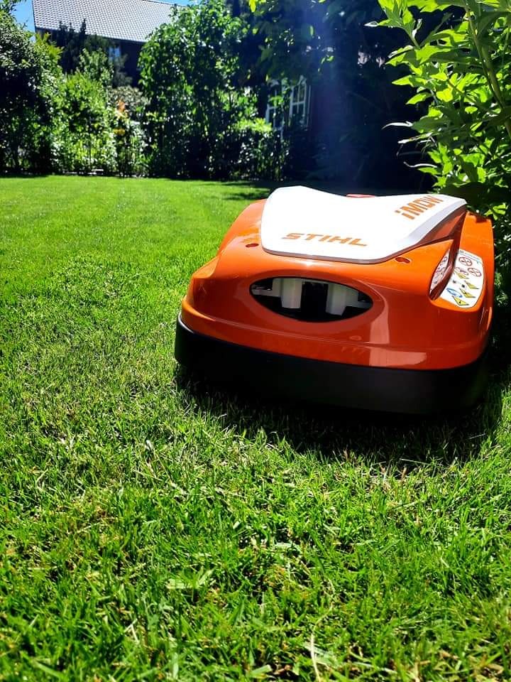 Pomarańczowy robot koszący Stihl iMOW! na zielonym trawniku przed domem w słoneczny dzień.