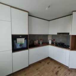 Nowoczesna kuchnia z białymi frontami i drewnianymi akcentami, widok na narożną zabudowę z piekarnikiem i płytą grzewczą, na blacie butelki płynów i akcesoria kuchenne, brązowa podłoga.